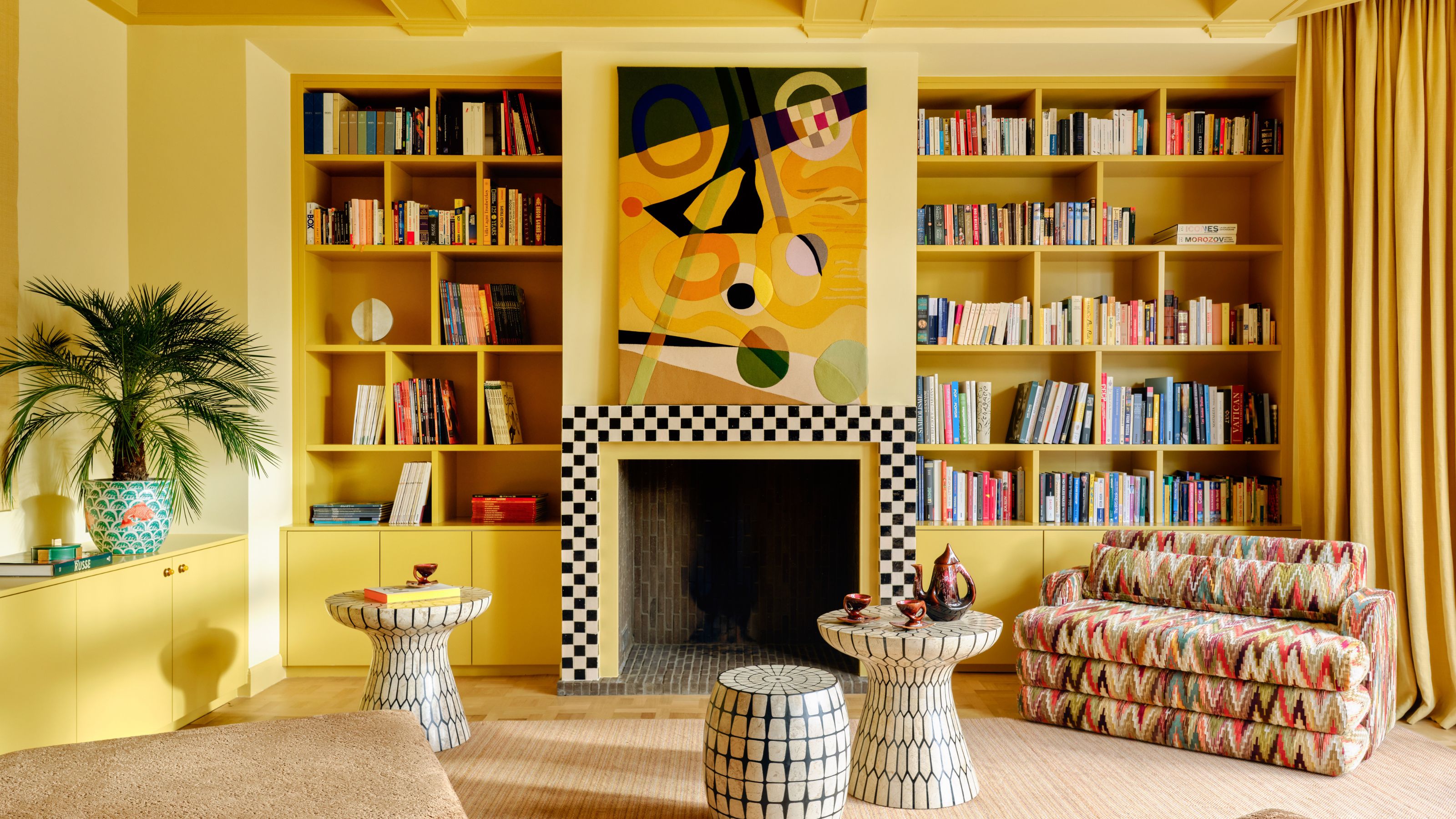 ein leuchtend gelb gestrichenes Wohnzimmer mit einer schachbrettartig gefliesten Feuerumrandung, Bücherregalen sowie vielseitigen Möbeln und Kunstwerken