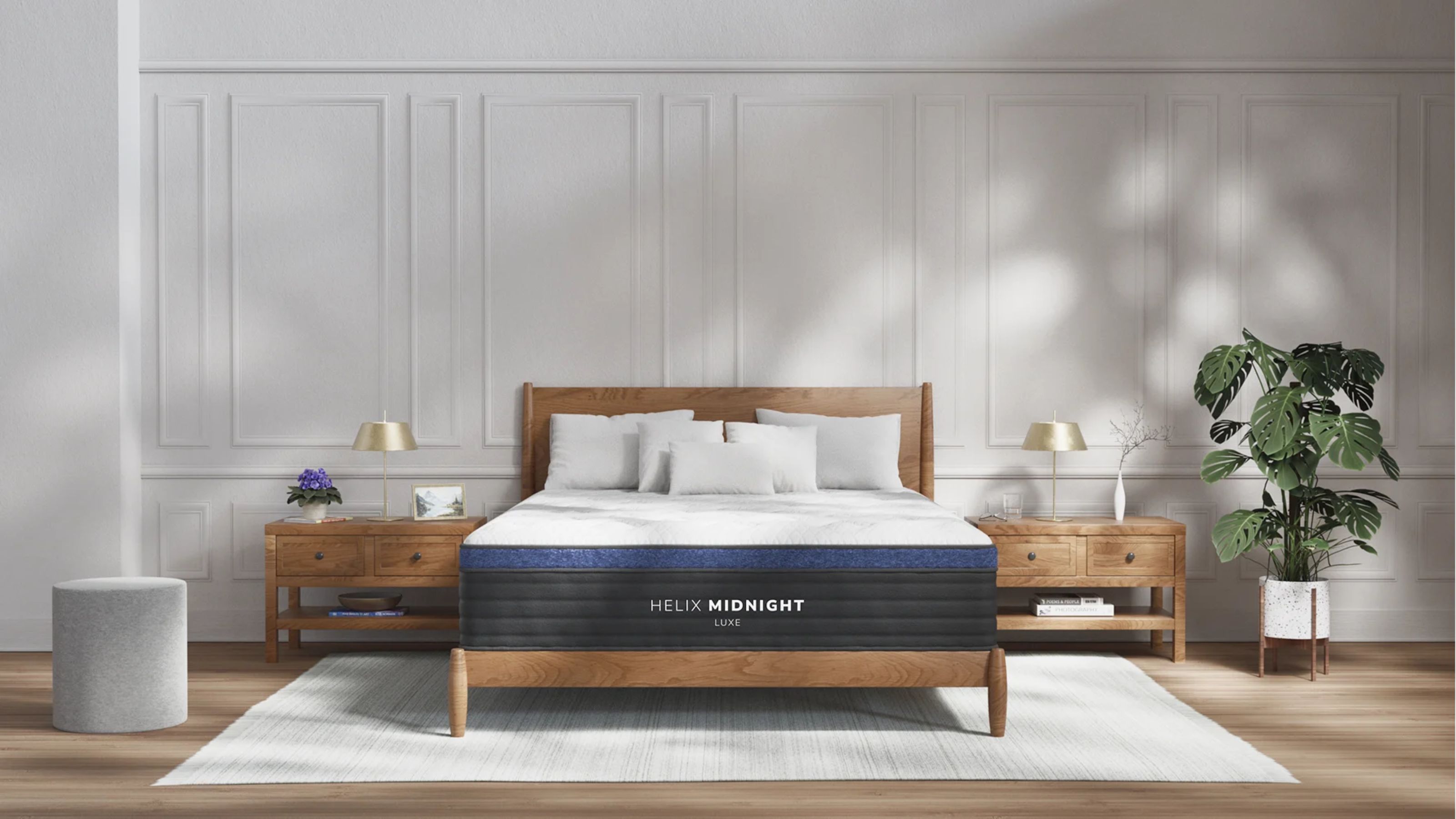 Helix Midnight Luxe Matratze auf einem Holzbettgestell in einem großen Schlafzimmer mit weiß getäfelten Wänden, Holzboden, einem hellgrauen Teppich und einer großen Zimmerpflanze sowie zwei passenden Nachttischen aus Holz.