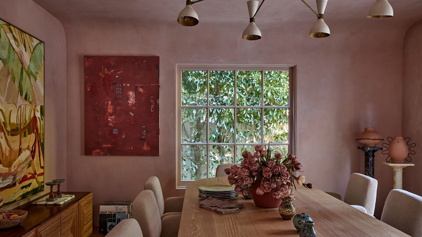 Rosafarbene Putzwände, ein langer Esstisch aus Eichenholz, skulpturale Beleuchtung, zeitgenössische Kunst und ein üppiger Blick auf den Garten schaffen ein warmes, vielschichtiges Esszimmer im modernen, organischen Stil.