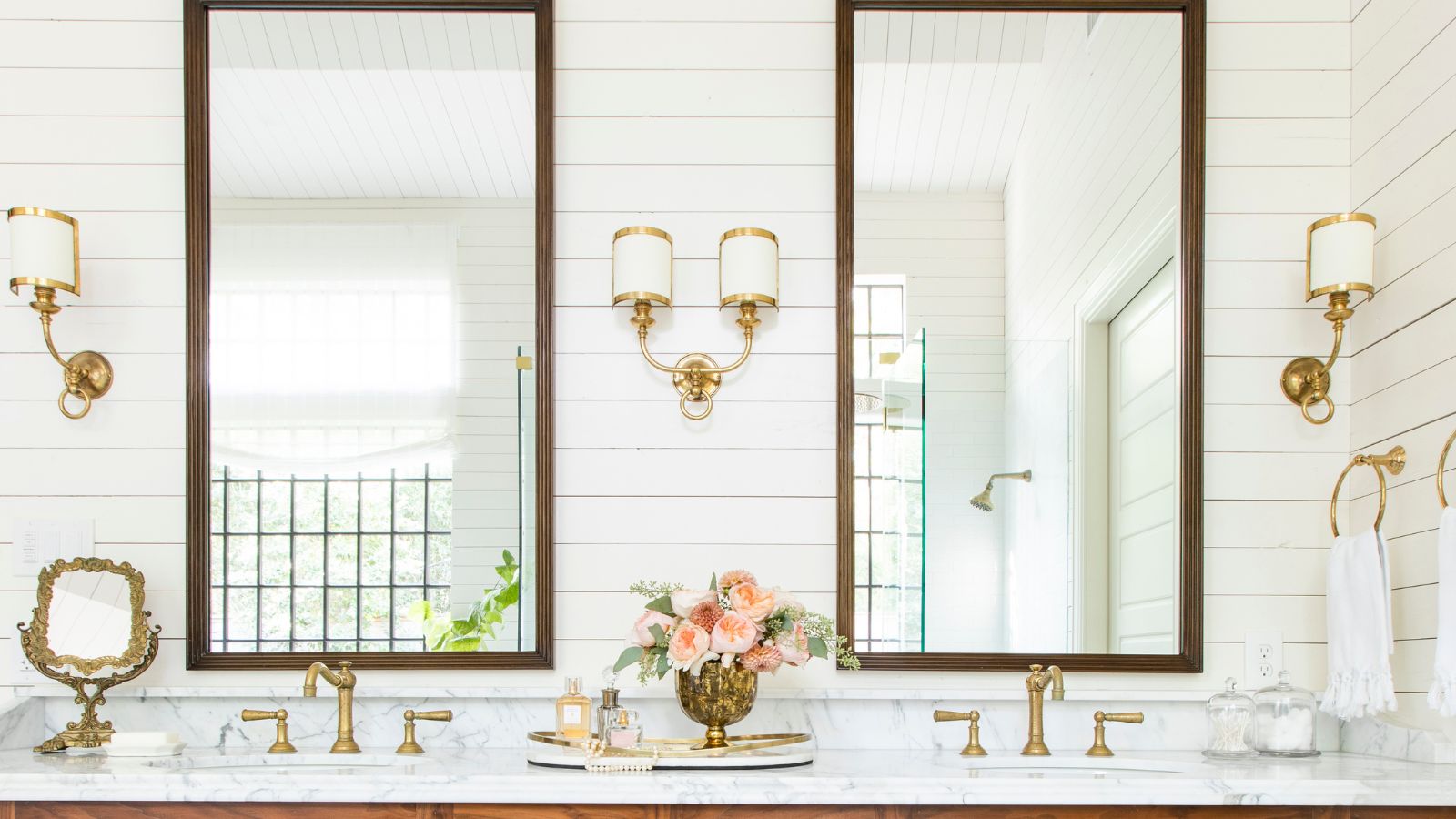 Badezimmer mit wolkenweiß gestrichenen getäfelten Wänden, großen Spiegeln mit Holzrahmen, einem Marmorwaschtisch mit Doppelwaschbecken und goldenen Wasserhähnen sowie goldenen und weißen Wandleuchten neben den Spiegeln