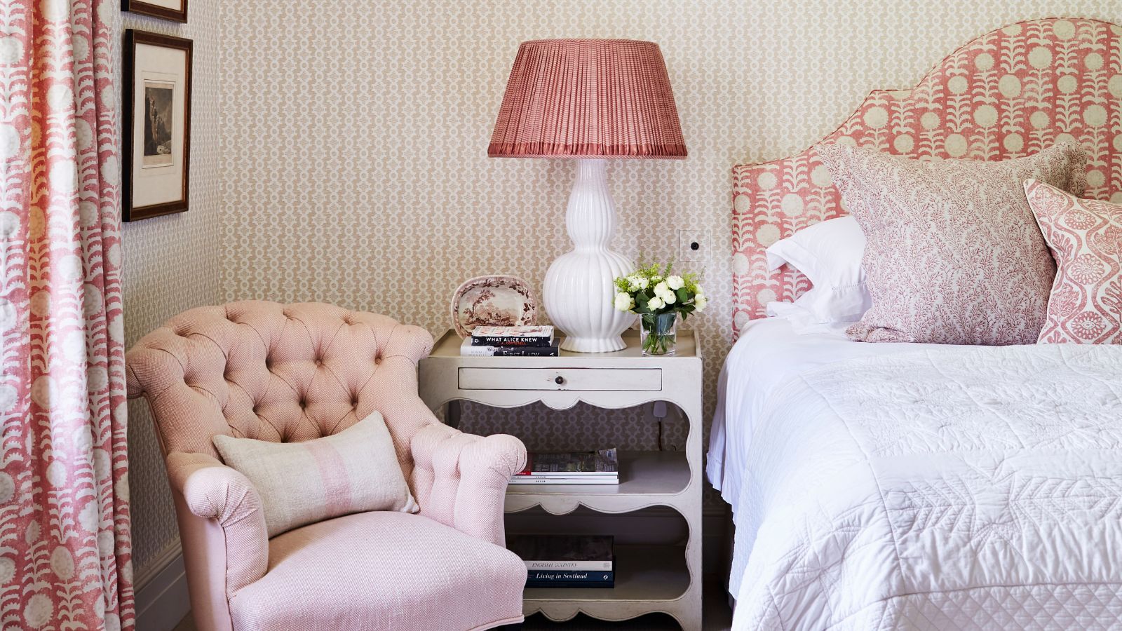 Ein weiß-rosa Schlafzimmer mit gemusterter Tapete, gemustertem Kopfteil, weißer Bettdecke, weißer Lampe mit rosa Lampenschirm, weißem Nachttisch, rosa Stuhl, gemusterten Vorhängen und gerahmten Kunstwerken.