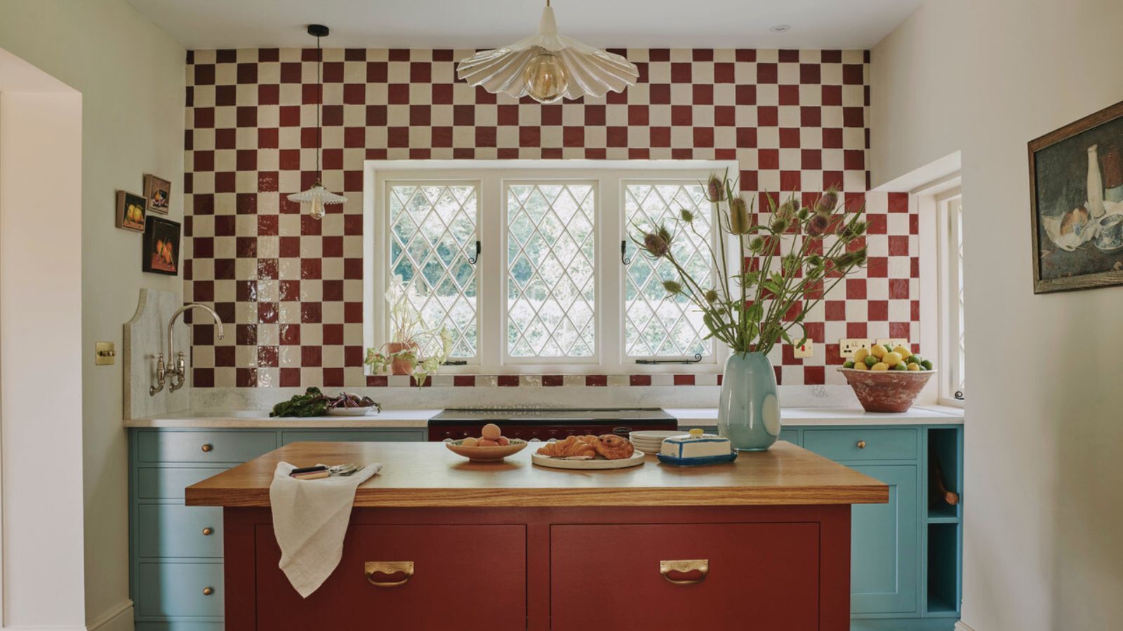 a kitchen with a checkerboard tile backsplash, lower blue painted cabinets, an island with a wooden top surface and red painted drawers, and a vase of flowers on the island.
