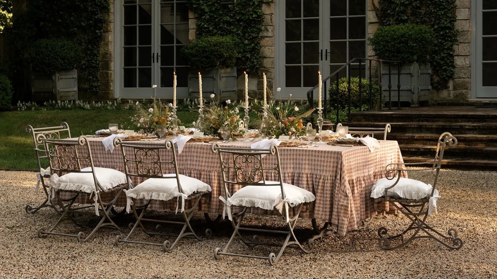 Großer langer Tisch im Freien auf einer schindelgedeckten Terrasse mit rosa karierter Rüschentischdecke, verzierten Stühlen, gedeckt mit Blumen und hohen Spitzkerzen