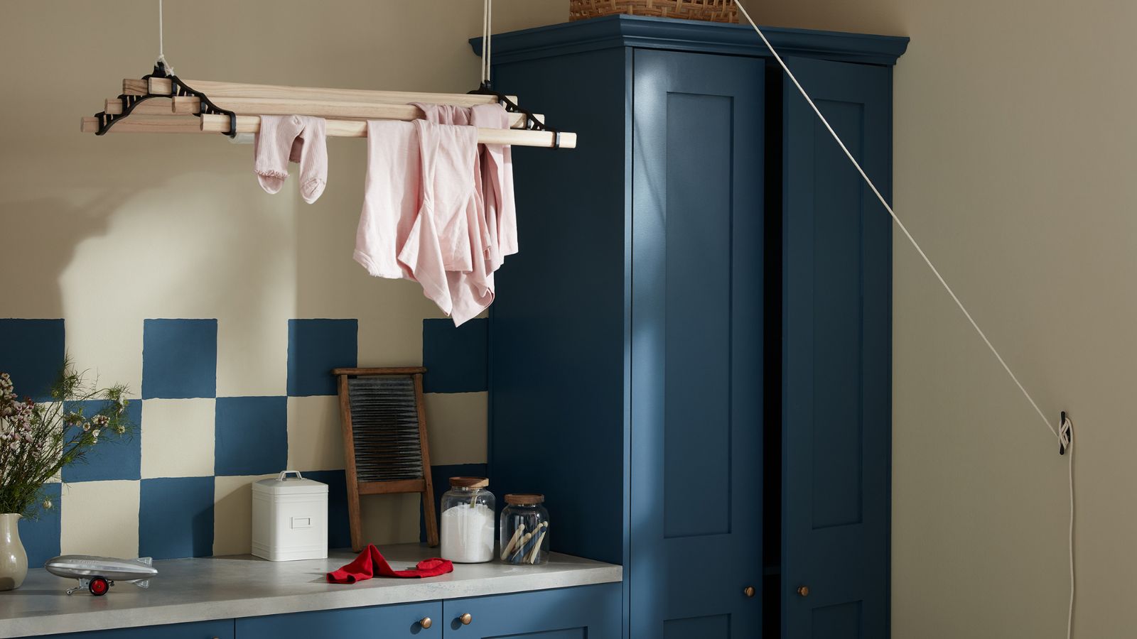 Dunkelblau gestrichene Schränke mit passenden blauen und weißen Fliesen in einer kleinen Waschküche. Darüber ein an der Decke hängender Wäscheständer, darauf ein paar rosa Kleidungsstücke. 