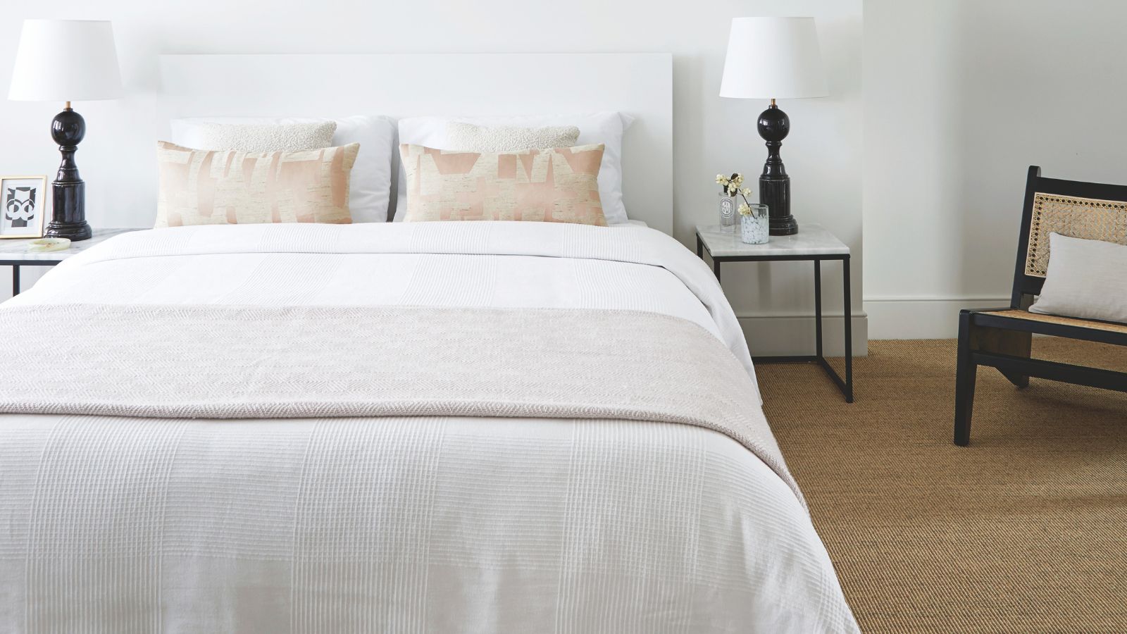 Weißes Schlafzimmer mit weißen Wänden, weißem Bett mit weißer Bettwäsche, Juteteppich, zwei passenden schwarzen Lampen mit weißen Schirmen auf beiden Seiten des Bettes und einem Stuhl aus Schwarz und Rattan mit weißem Kissen.