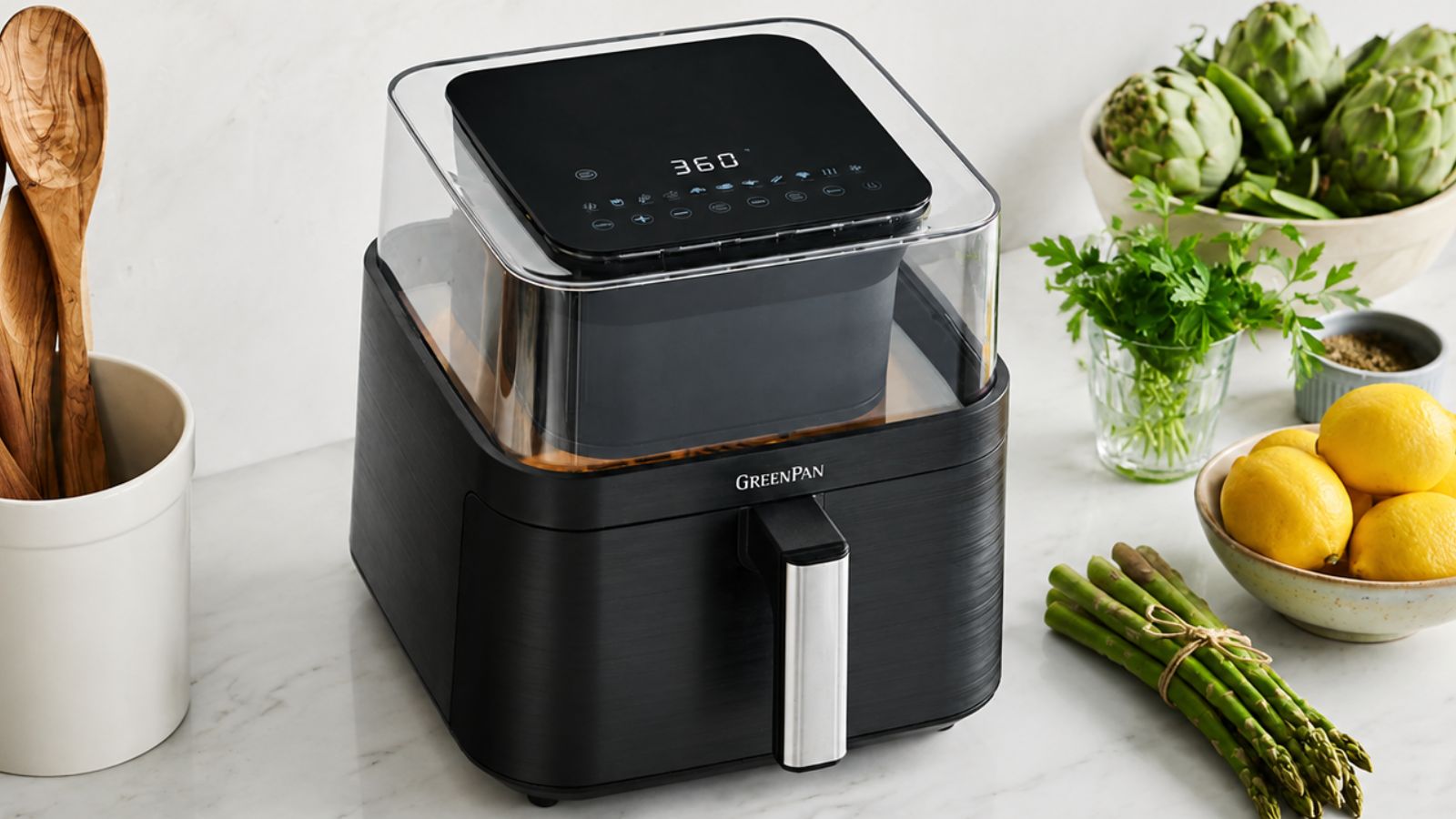 Die GreenPan Bistro 360 Clear View Heißluftfritteuse auf einer Küchentheke aus Marmor neben einem Topf mit Holzutensilien und etwas frischem Frühlingsgemüse. 