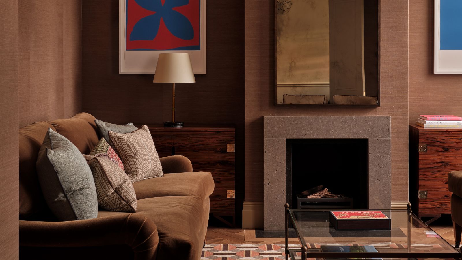 Wohnzimmer mit braunen Grasstoffwänden, cremefarbener Decke, Laternen-Pendelleuchte, geometrischem Teppich in Orange und Braun, Couchtisch aus Glas, braunem Samtsofa und einem Kamin mit angelaufenem Spiegel darüber