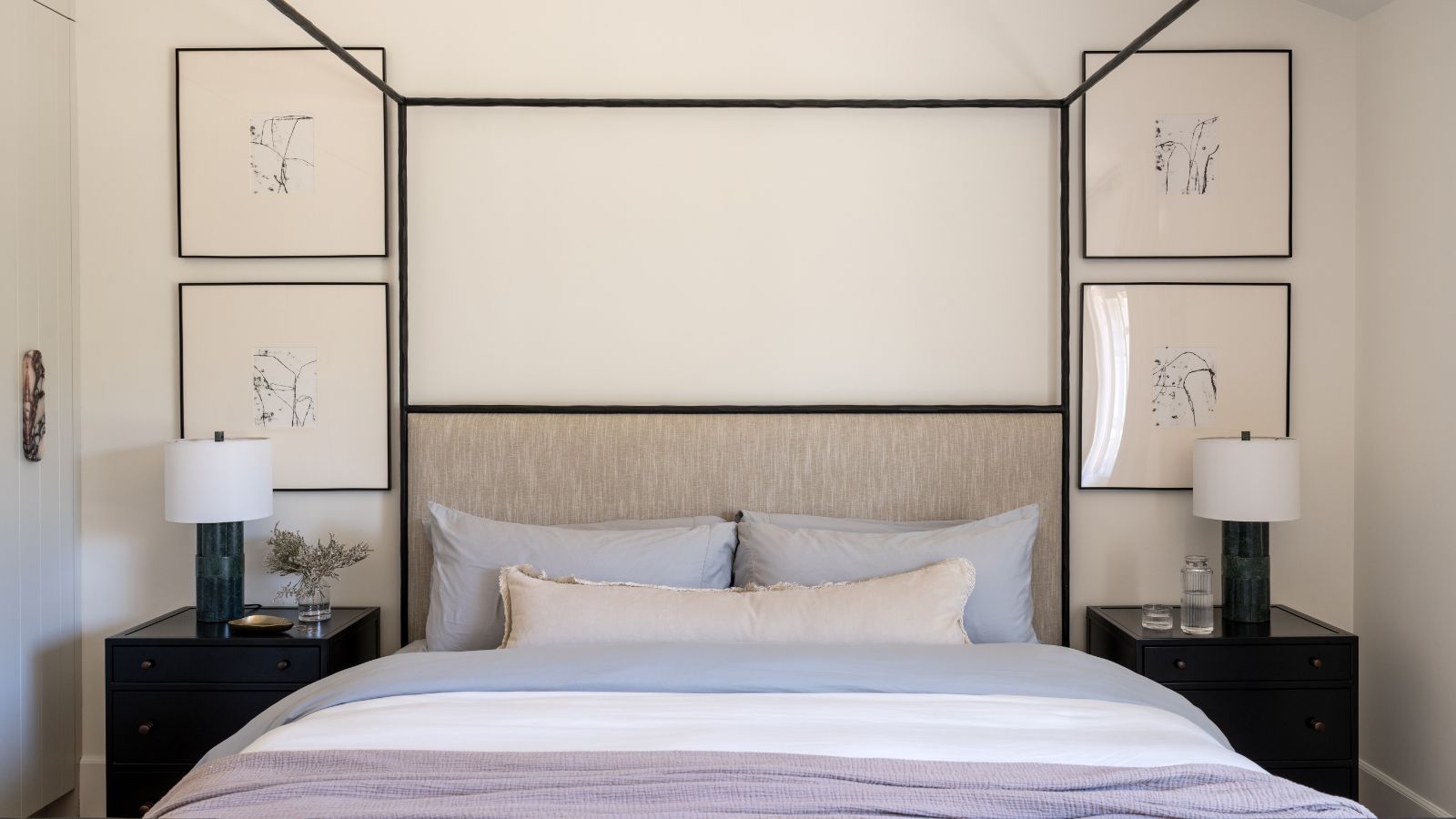 Schlafzimmer mit einem schwarzen Himmelbett aus Metall, lila Leinenbettwäsche und einem neutralen Lendenkissen, vier gerahmten Drucken an der Wand in schwarzen Rahmen und schwarzen Nachttischen auf beiden Seiten mit Tischlampen.
