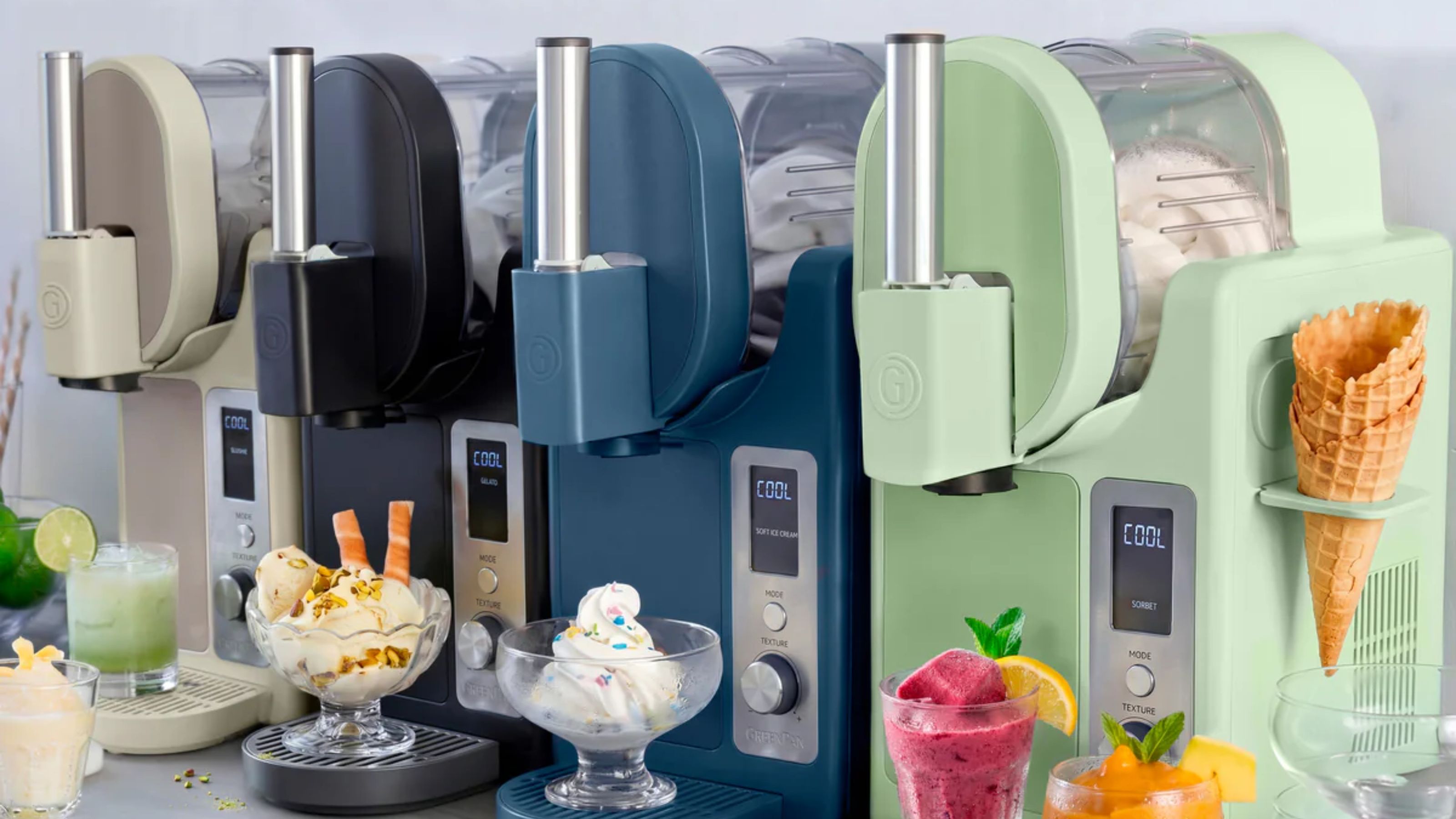 Die Eismaschine GreenPan Frost Pro in den Farben Grün, Blau, Schwarz und Beige steht aufgereiht auf einer Theke, darunter befinden sich Eisbecher in Schüsseln. 