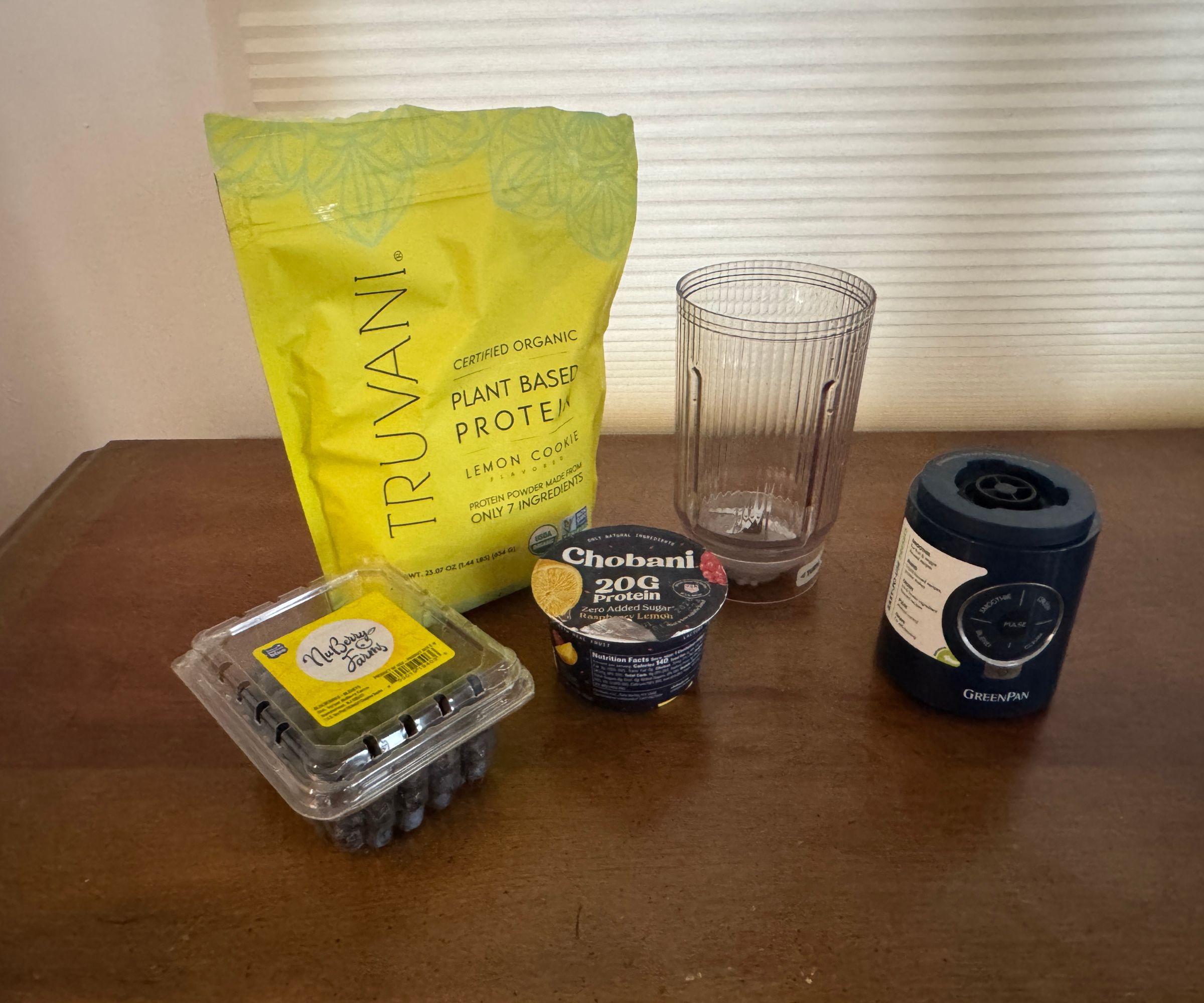 Eine Tüte Proteinpulver, ein Joghurt und einige Blaubeeren neben einem kleinen tragbaren Mixer.
