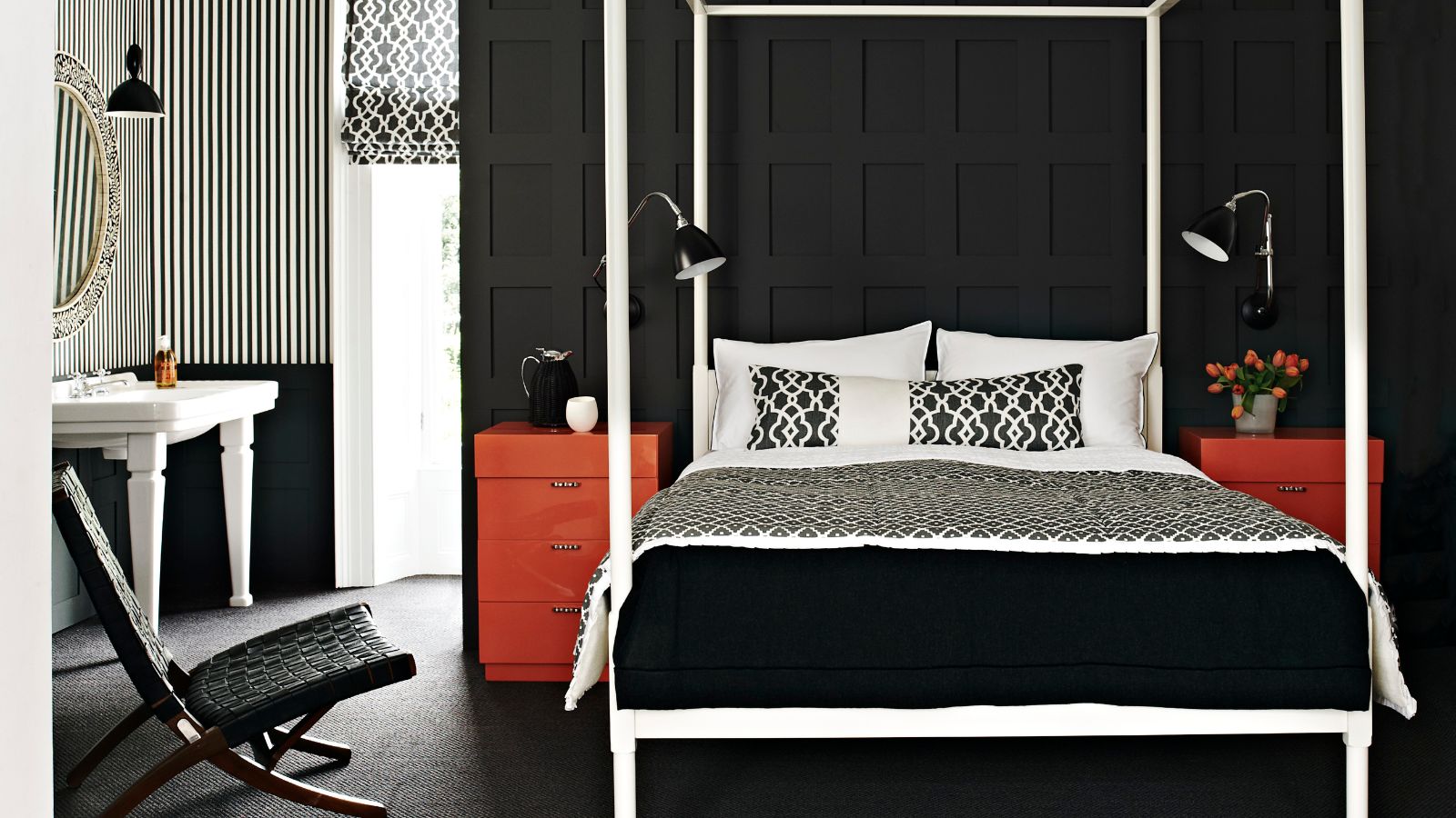 Ein schwarzes Schlafzimmer mit Wandvertäfelung, einem großen weißen Himmelbett und zwei roten Kommoden auf beiden Seiten mit passenden schwarzen Lampen. Auf der linken Seite befindet sich eine schwarz-weiß gestreifte Wand, ein weißes Waschbecken, ein großes Fenster, ein runder Spiegel und ein schwarzer Stuhl.