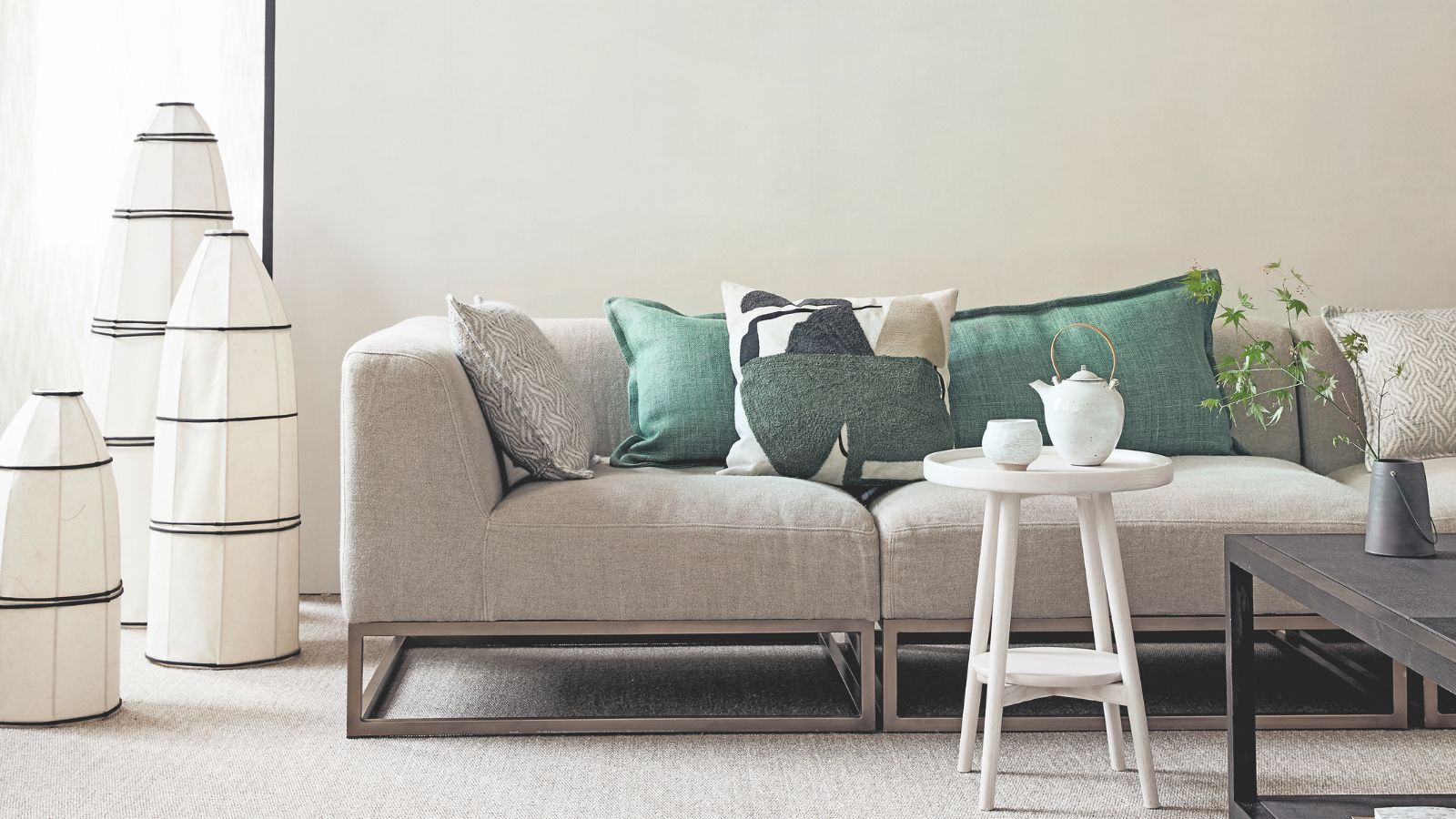 Ein weißes Wohnzimmer mit einer hellgrauen Couch, grünen Kissen, einem weißen Tisch mit weißem Teeservice, einem schwarzen Couchtisch mit grüner Pflanze und drei großen schwarzen und weißen Lampen neben der Couch.