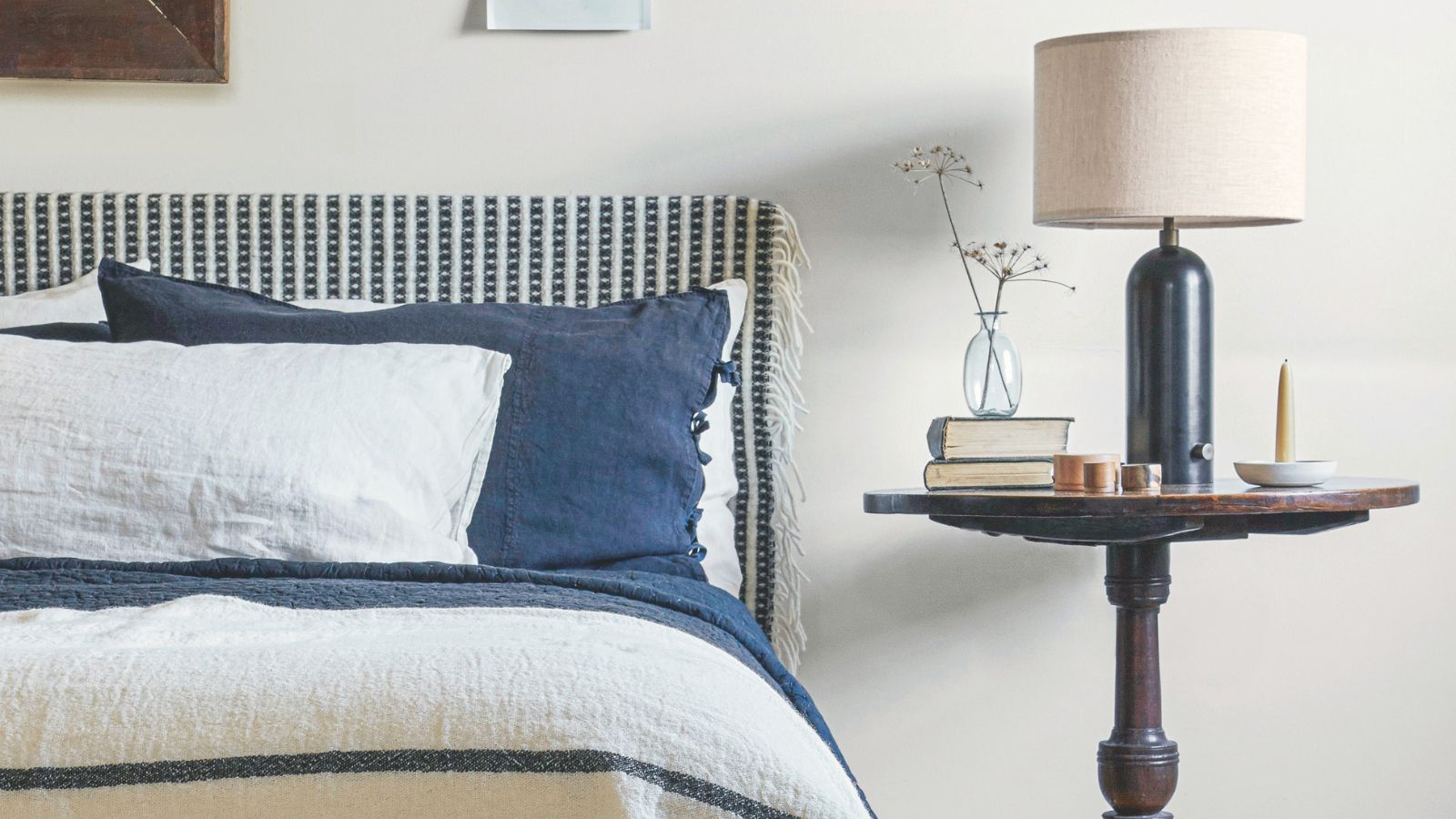 Neutrales Schlafzimmer mit weißen Wänden und einem blau-weiß gemusterten Bett mit passender Bettwäsche und Kissen. Daneben ein runder Nachttisch aus dunklem Holz, eine dunkelblaue Lampe mit weißem Schirm und eine Glasvase.