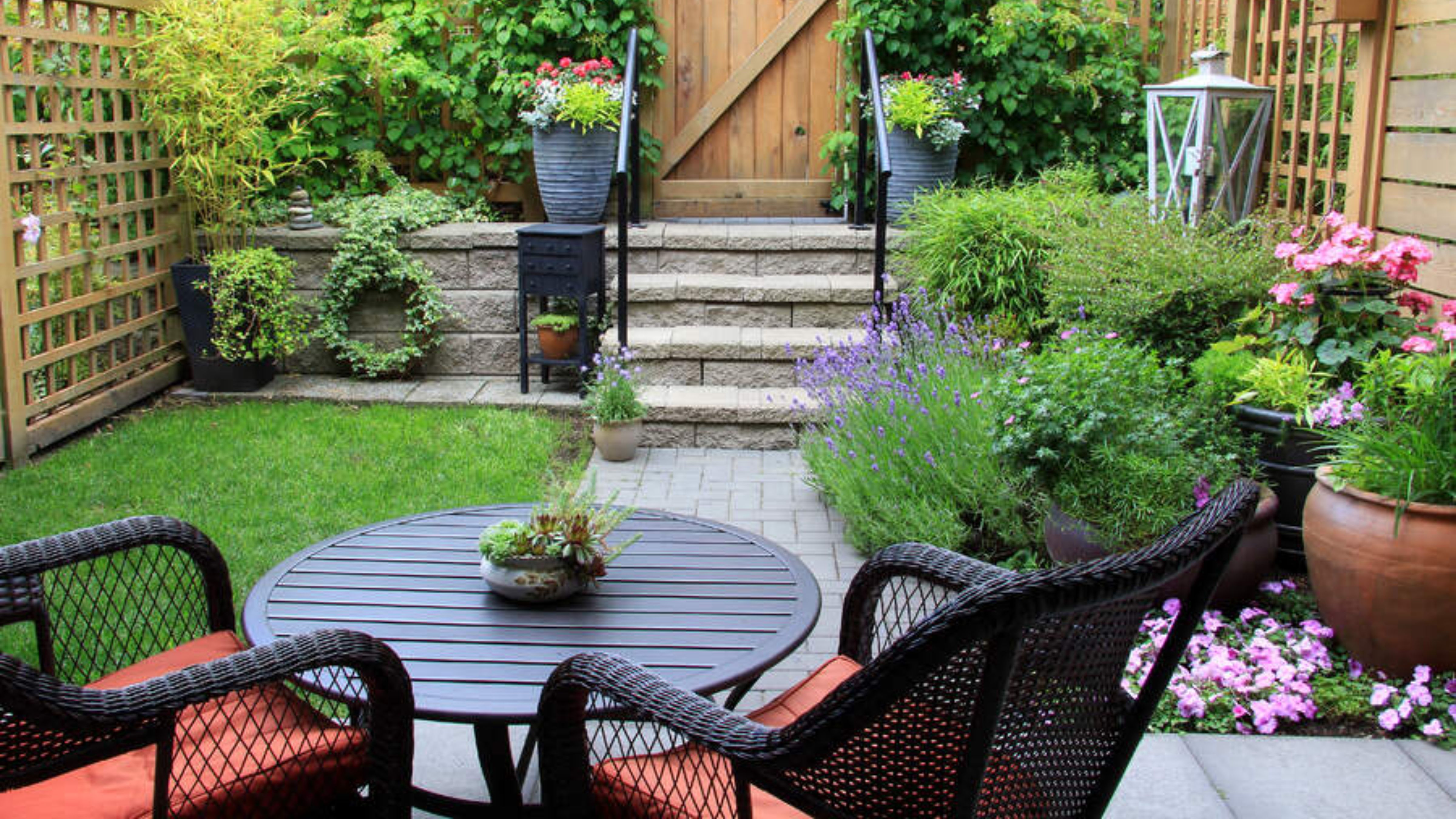 Kleiner Hinterhofbereich mit kleinem Garten, Steinstufen und einem schwarz gewebten Bistro-Set mit ziegelroten Kissen.