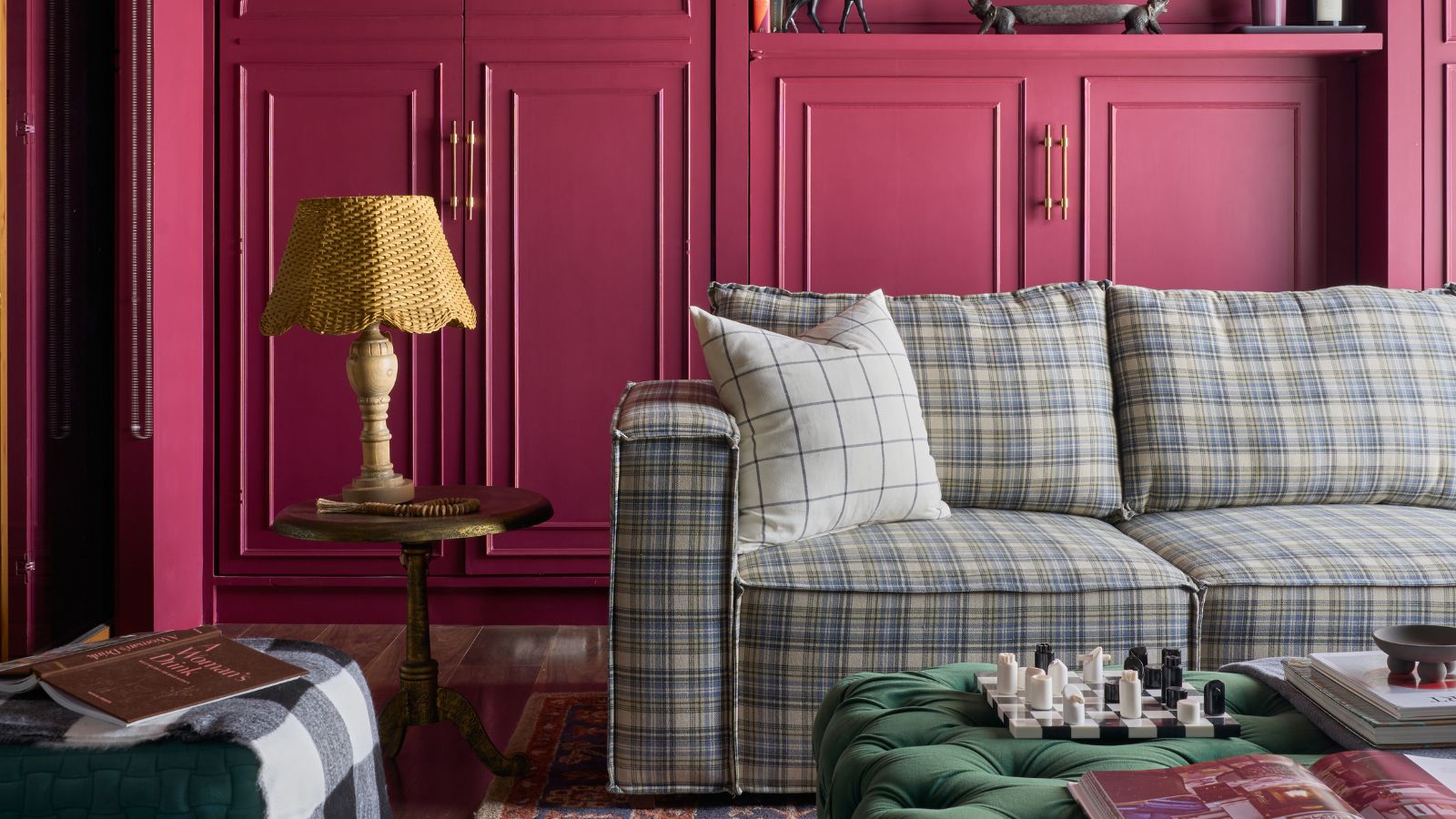 Wohnzimmer mit himbeerrosa Schränken, kariertem blauem Sofagarnitur, Perserteppich, grünem Samthocker, Beistelltisch mit Lampe und Pflanze auf dem Boden