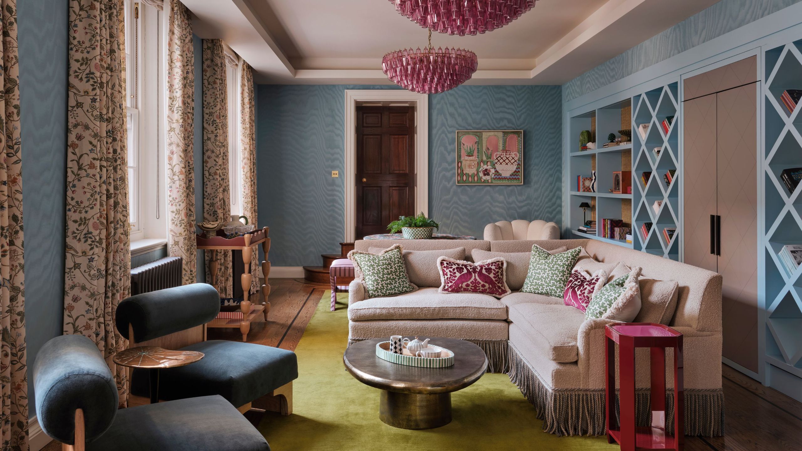 a blue wallpapered home library with a long wall of bookcases, a corner settee with fringing, pink chandeliers and side table