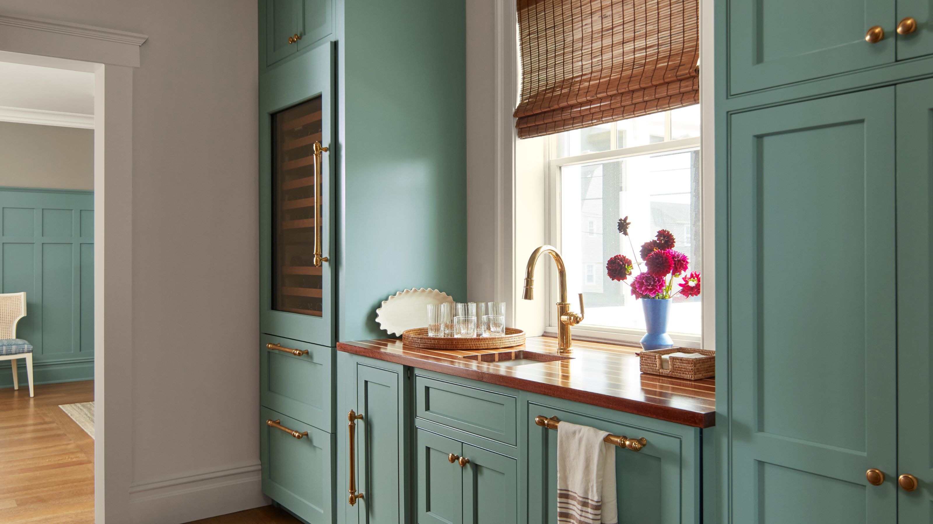 Küche mit in Benjamin Moores Stratton Blue gestrichenen Möbeln, einer Holzarbeitsplatte mit goldenem Wasserhahn, einem Rattanrollo über dem Fenster und einer Vase mit rosa Blumen auf der Fensterbank.