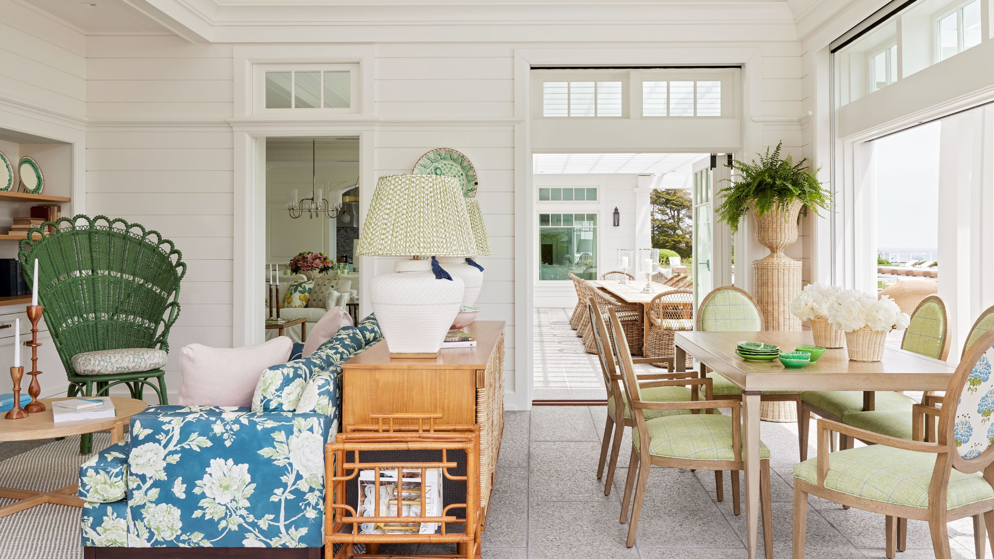 Traditionelles Küstenhaus mit weißen Schiffswänden, großen Fenstern, einem Esstisch mit grünen Stühlen, einem Sofa mit Blumenmuster und einer Holzkonsole und zwei Lampen dahinter sowie einem grünen Pfauenstuhl in der Ecke.