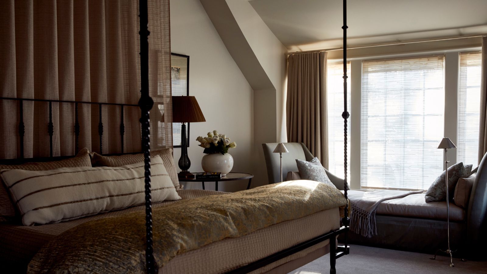 A cozy neutral bedroom with warm white walls, a large window with a bench seat and beige curtains, carpeted floor, a four-poster canopy bed with layered neutral bedding.