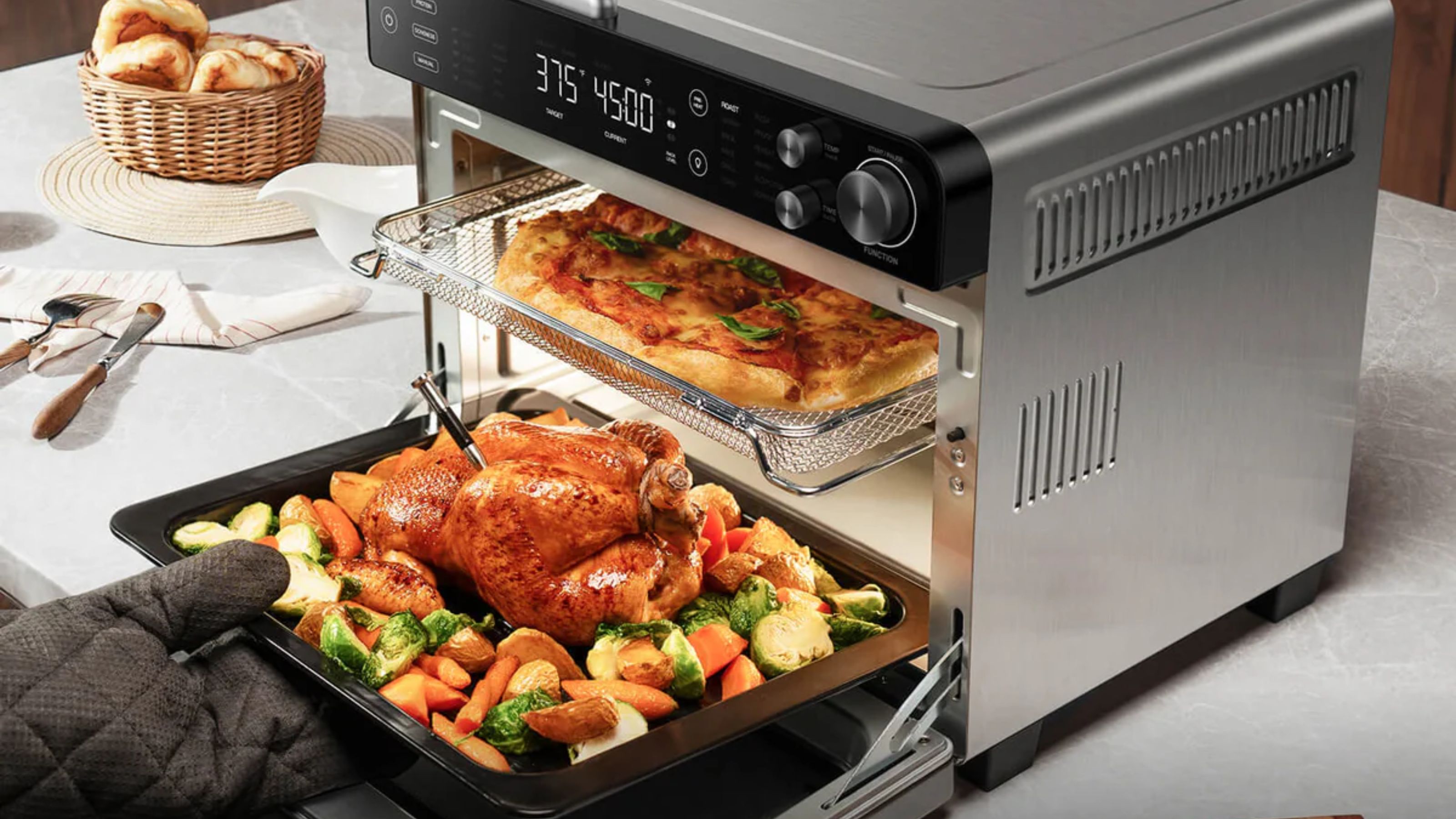 Ein Typhur-Synchronluftfritteusenofen auf einer weißen Arbeitsplatte, mit einer Pizza auf dem oberen Regal und jemandem, der ein Brathähnchen auf dem unteren Regal herausholt. 