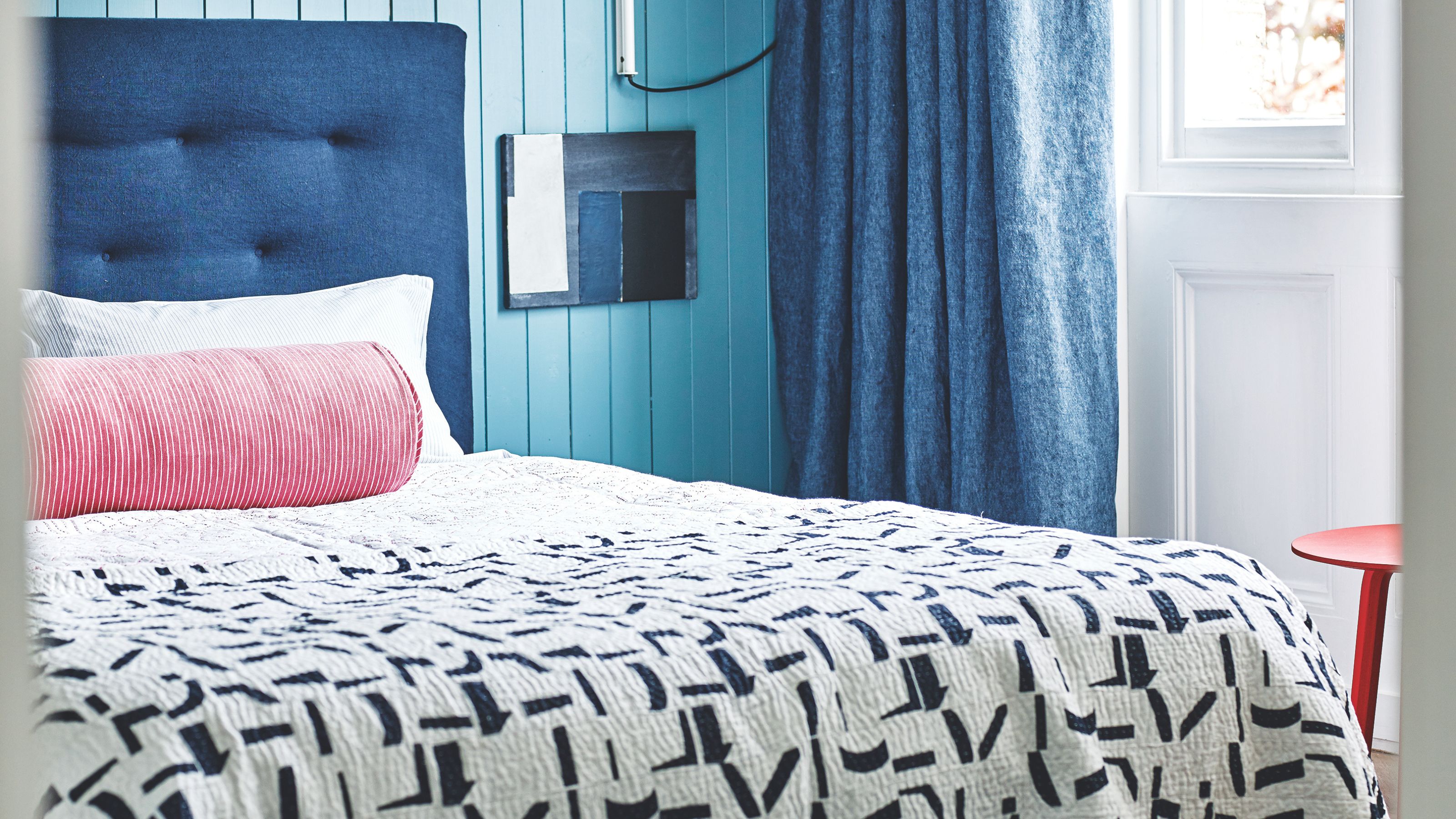 Blau getäfeltes Schlafzimmer mit blauem Doppelbett mit weißer Bettwäsche, rosa zylindrischem Kissen, gemusterter Steppdecke und großen weißen Fenstern mit dunkelblauen Vorhängen neben einem roten Hocker.