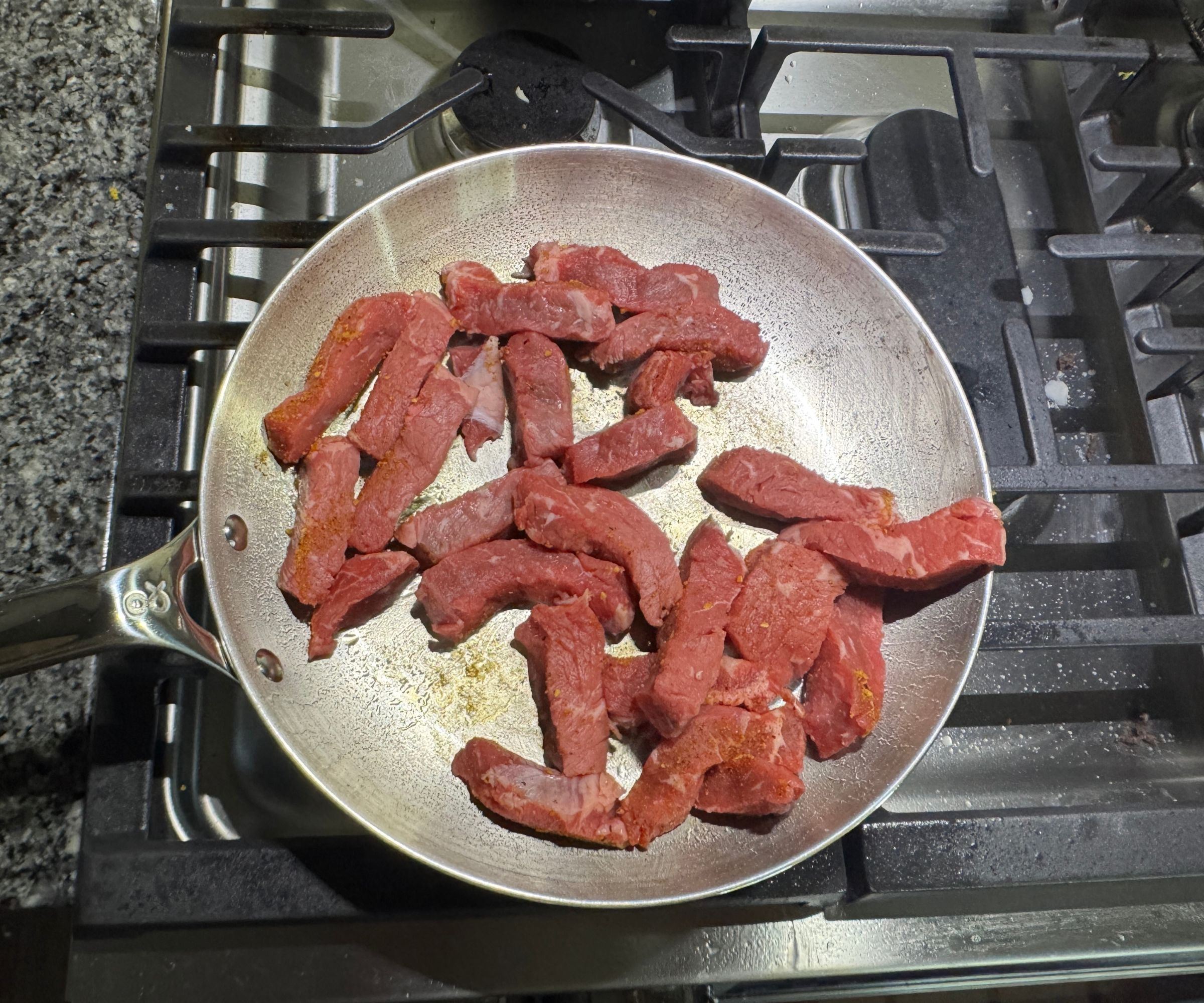 Eine kleine Bratpfanne aus Edelstahl auf einem Gasherd, gefüllt mit rohen Steakstreifen