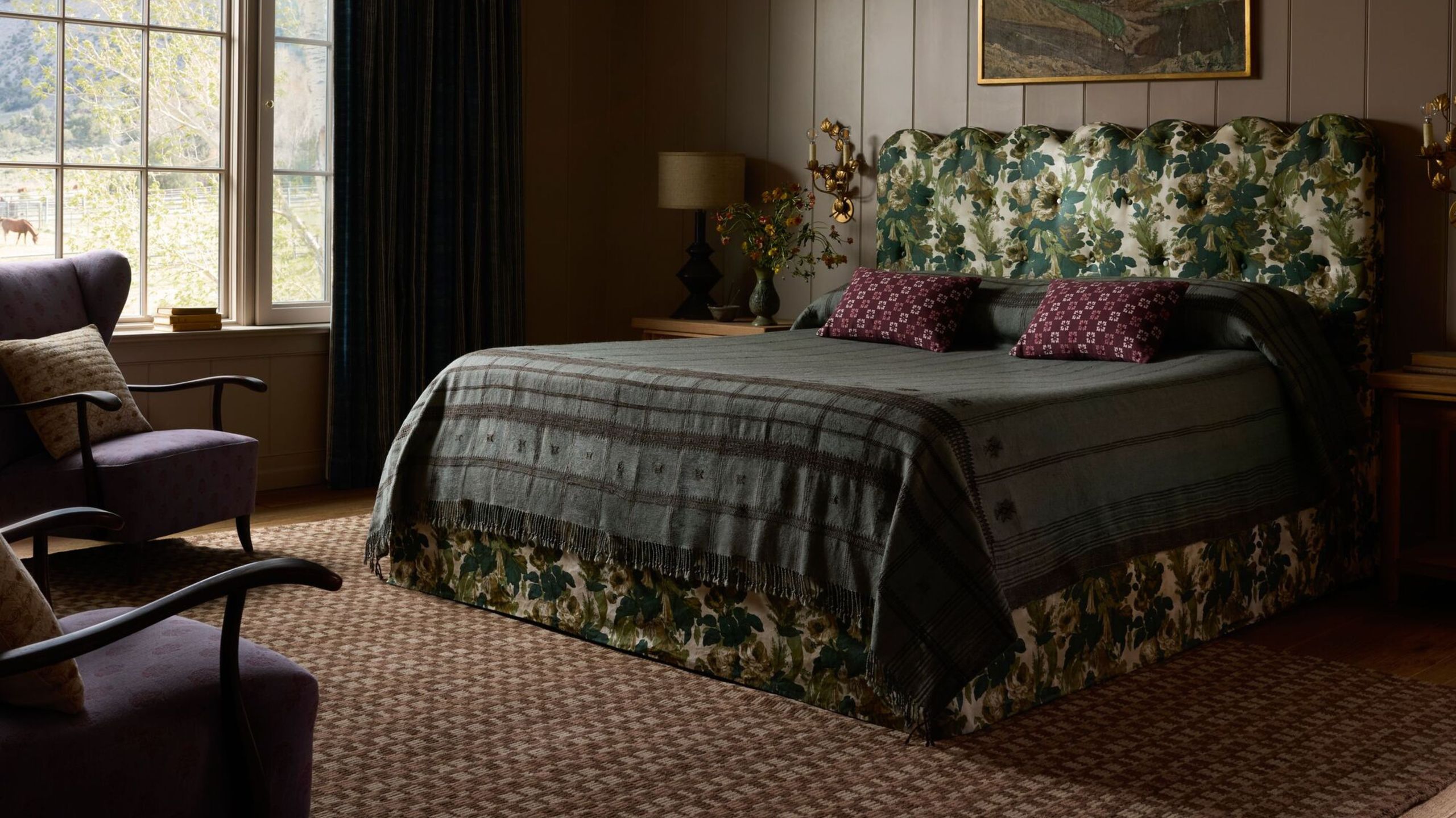 Moody, richly layered bedroom featuring a neutral heritage patterned rug, tapestry-print bed frame flanked by two ornately gilded candle sconces, and eclectic bedding.