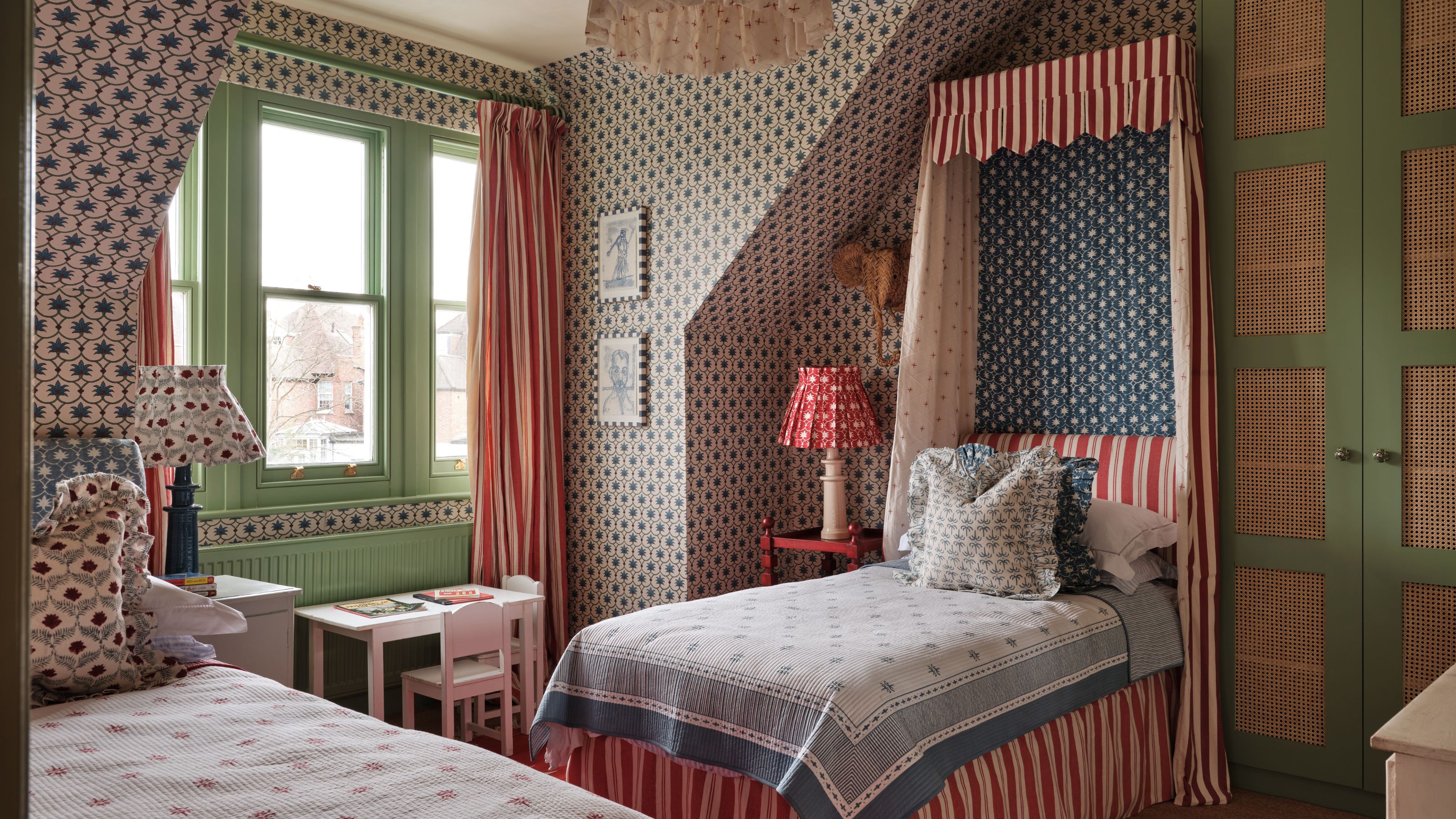 Schlafzimmer mit gemusterter Tapete, einem grünen Fensterrahmen, einem gestreiften Himmelbett mit blau-weißer Bettwäsche, einem kleinen Schreibtisch unter dem Fenster und einem weiteren Einzelbett gegenüber mit einem Blumenkissen.