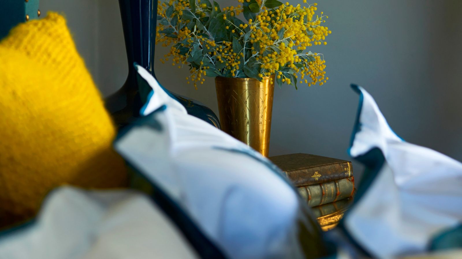 Blaues Schlafzimmer mit weißen Kissen, goldenen Kissen und einer goldenen Vase voller gelber Blumen mit grünen Blättern.