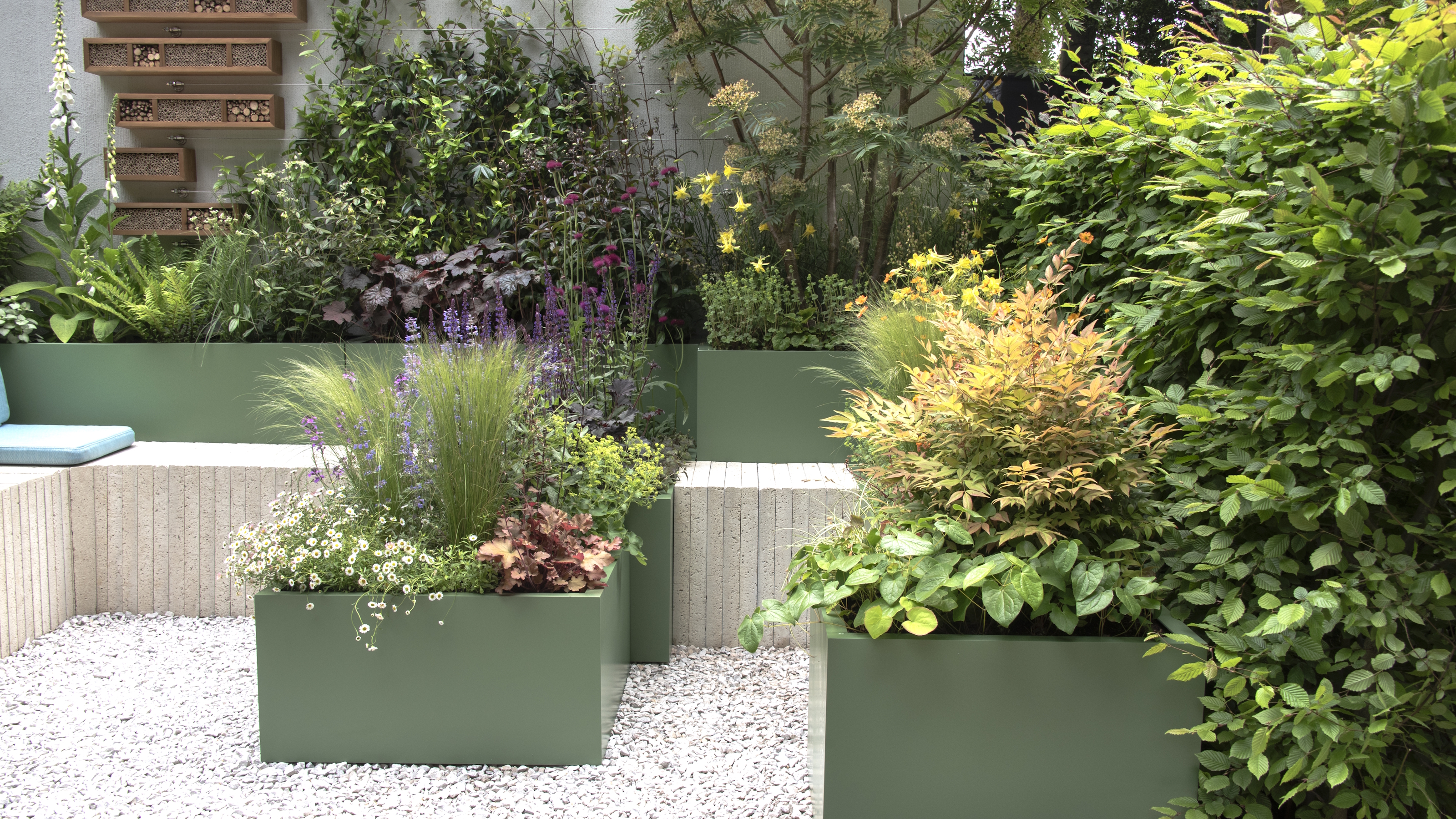 Pflanzen, die in grünen Behältern und Hochbeeten wachsen, umgeben einen Sitzbereich und eine mit Kies bestreute Terrasse auf der Chelsea Flower Show