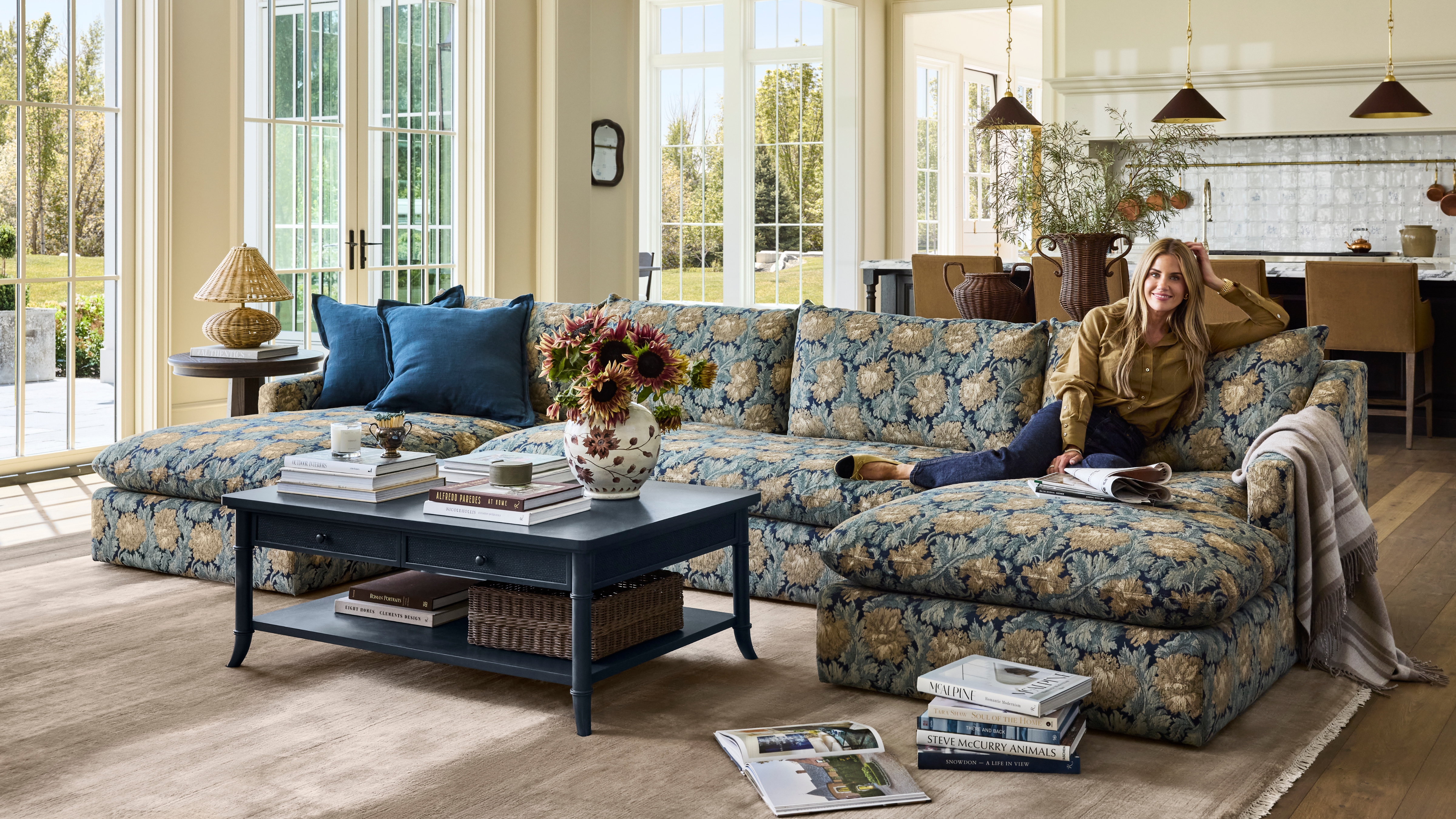 Shea McGee lächelt auf einem floralen Jacquard-Teil ihrer neuen McGee & Co. Herbstkollektion 2026, die mit einem marineblauen Couchtisch mit Stauraum, einem taupefarbenen Teppich, einer gewebten Tischlampe und Sammelobjekten ausgestattet ist