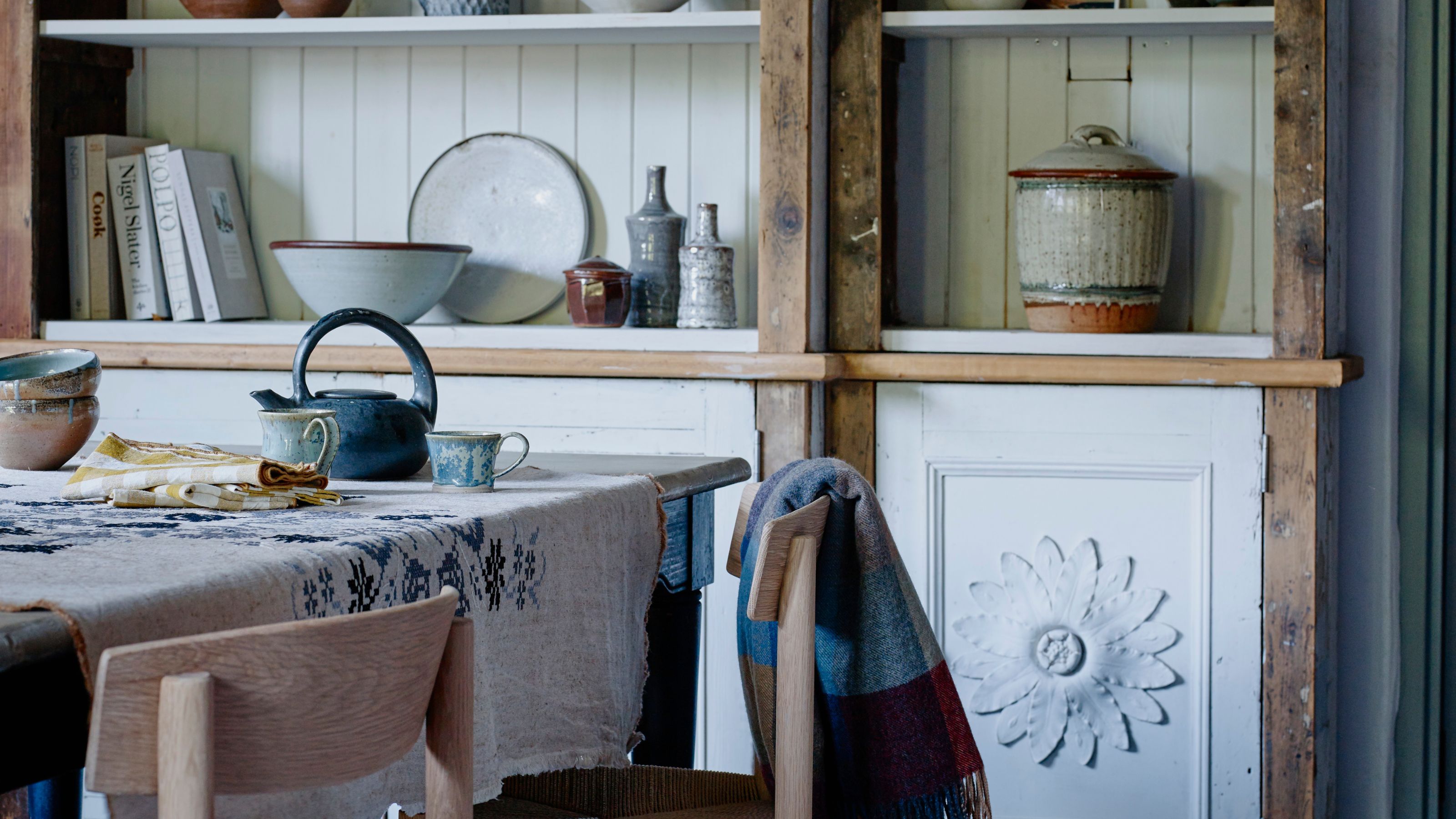 Esszimmer mit Esstisch mit Holzstühlen und Regal mit weißer Wandvertäfelung, Blumenmotiven sowie verschiedenen Keramikartikeln und Rezeptbüchern