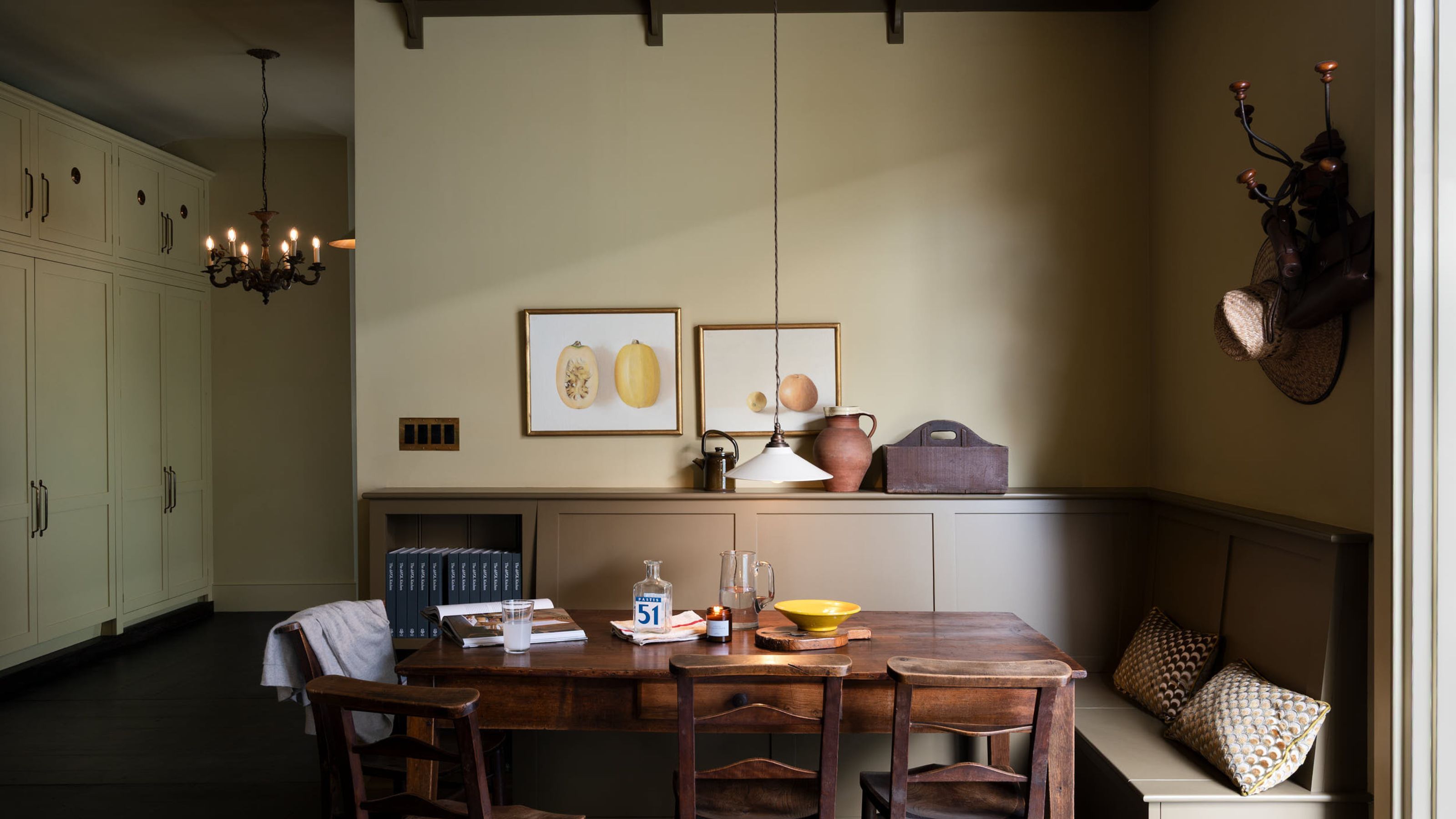 Essecke im Shaker-Stil mit salbeigrünen Wänden, Esstisch aus dunklem Holz, eingebauten Sitzbänken, rustikalen Keramiktöpfen, Stilllebengemälden mit Früchten an den Wänden und Holzbalken.