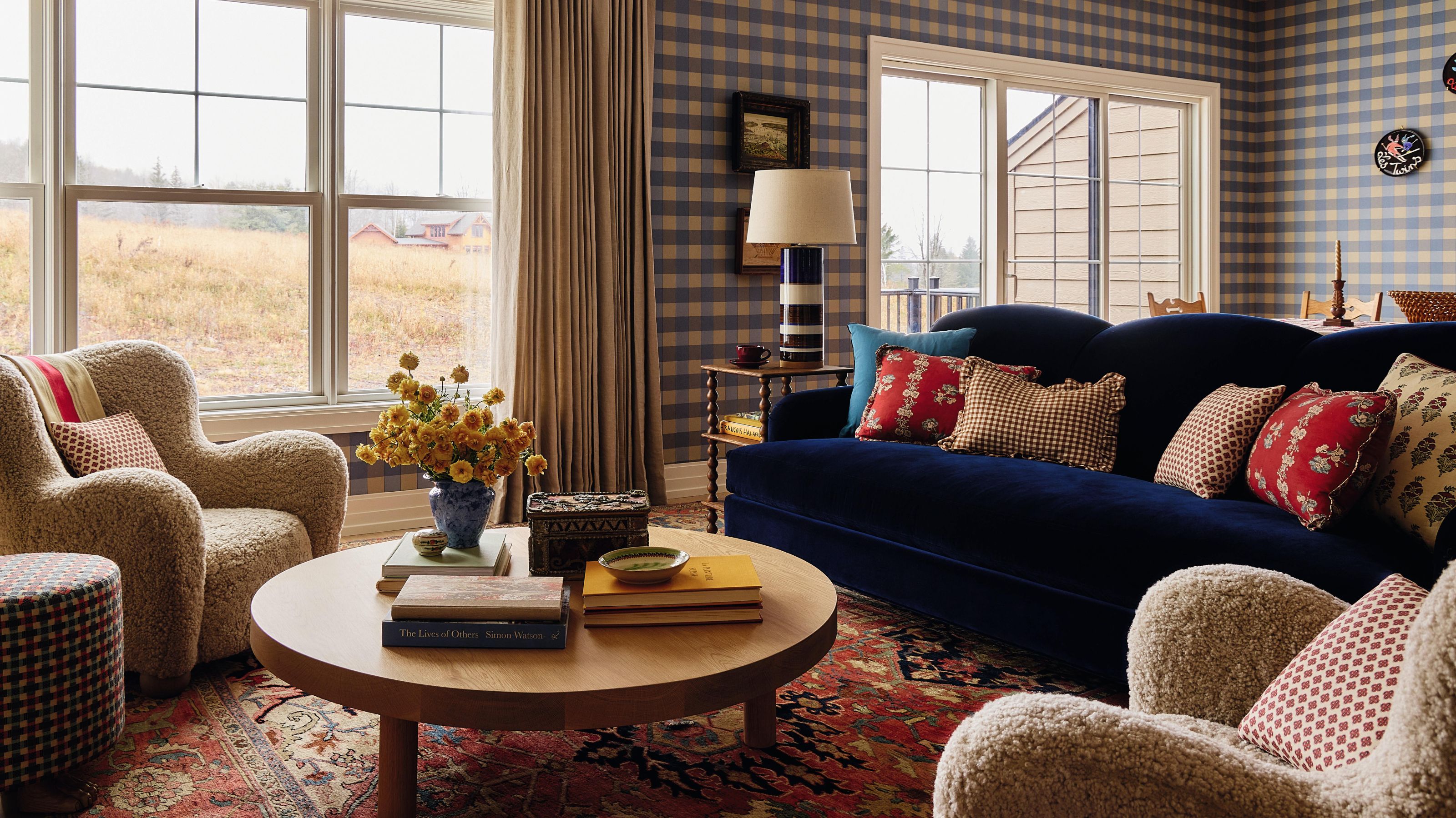 Ein offener Wohnbereich in einem Haus in den Catskills mit blauer Gingham-Tapete, einem marineblauen Samtsofa, einem roten Vintage-Teppich, Schaffellsesseln und einem runden Couchtisch aus Holz
