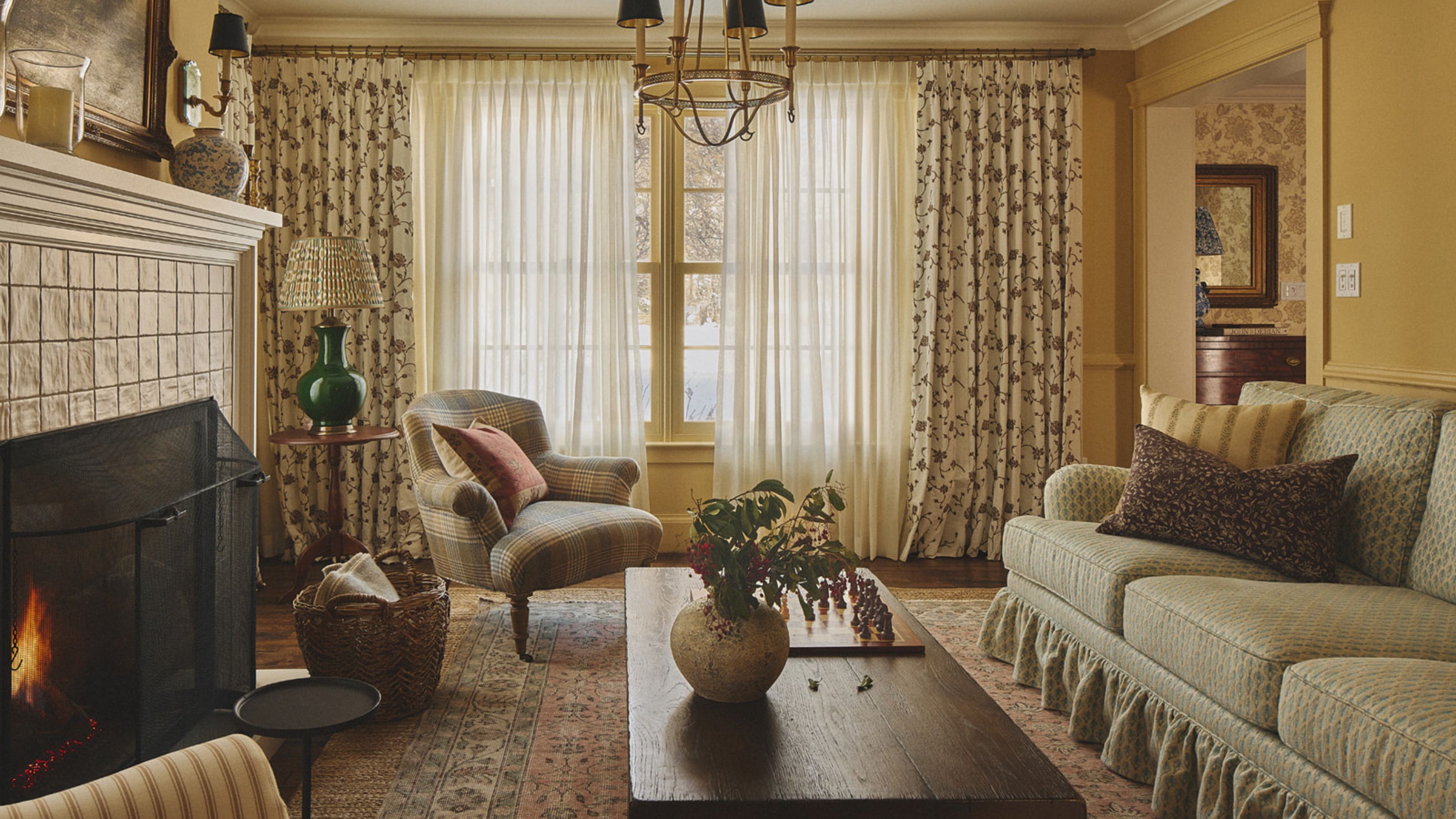 ein warmes gelbes Wohnzimmer mit geblümtem Sofa, geblümten Vorhängen, Vintage-Holzmöbeln und einem brennenden Kachelofen