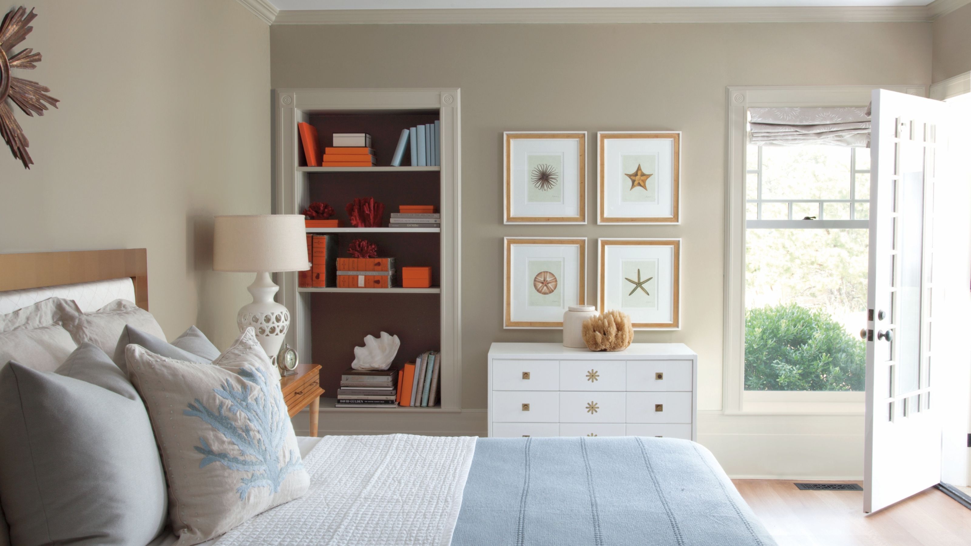 Küstenschlafzimmer mit Wänden von Pashmina by Benjamin Moore, einem Bett mit hellblauer und weißer Bettwäsche, einer Wand mit nautischen Drucken, einer weißen Kommode und eingebauten Bücherregalen mit orangefarbenen Kästen.