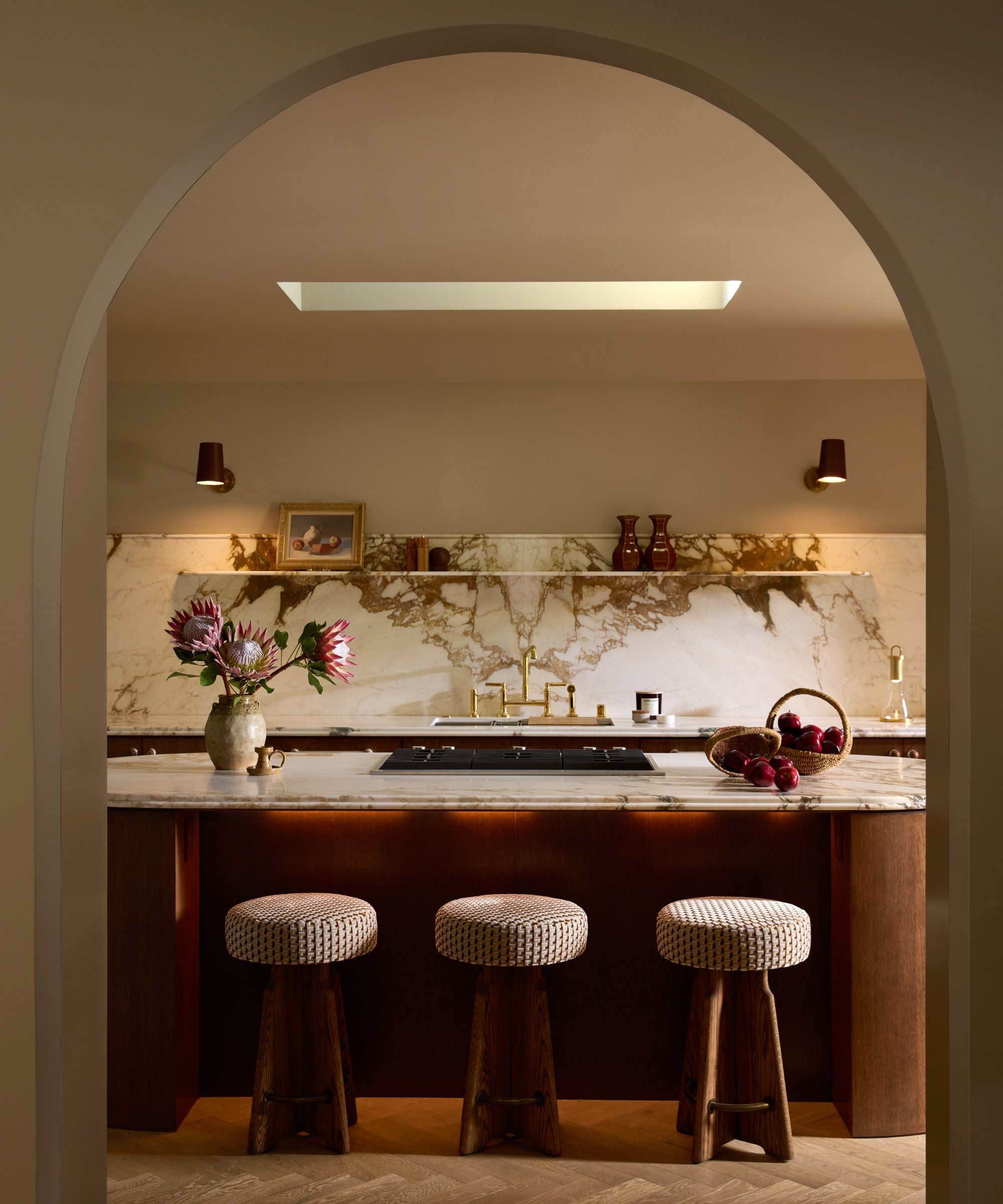 Bogentür mit Blick in die moderne Küche mit runder Kücheninsel aus Holz und Marmor mit drei runden Hockern, passender Marmorrückwand mit Regalen und zwei Wandleuchten