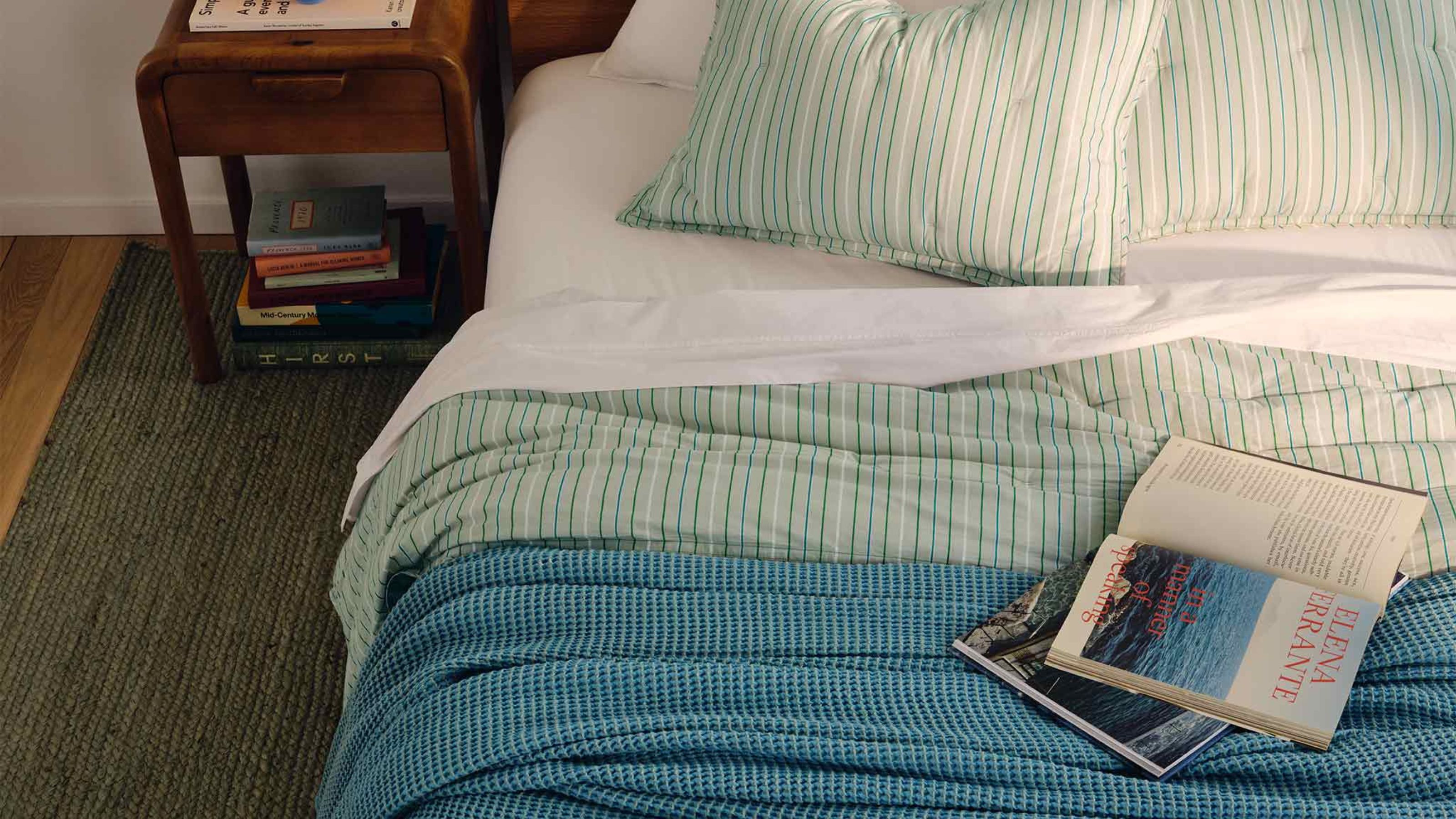 Waffeldecke aus blauer Baumwolle von Brooklinen auf einem Doppelbett mit gestreifter grüner Bettwäsche, daneben ein aufgeschlagenes Buch und ein Nachttisch aus dunklem Holz mit einem Stapel Bücher.