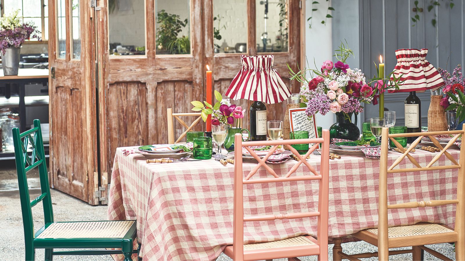 Rustikaler Tisch mit Gingham-Tischdecke, rustikalen bunten Holzstühlen, plissierten gestreiften Lampen, Stabkerzen, grünem Glas und Blumen, mit rustikalen, geretteten Türen im Hintergrund.