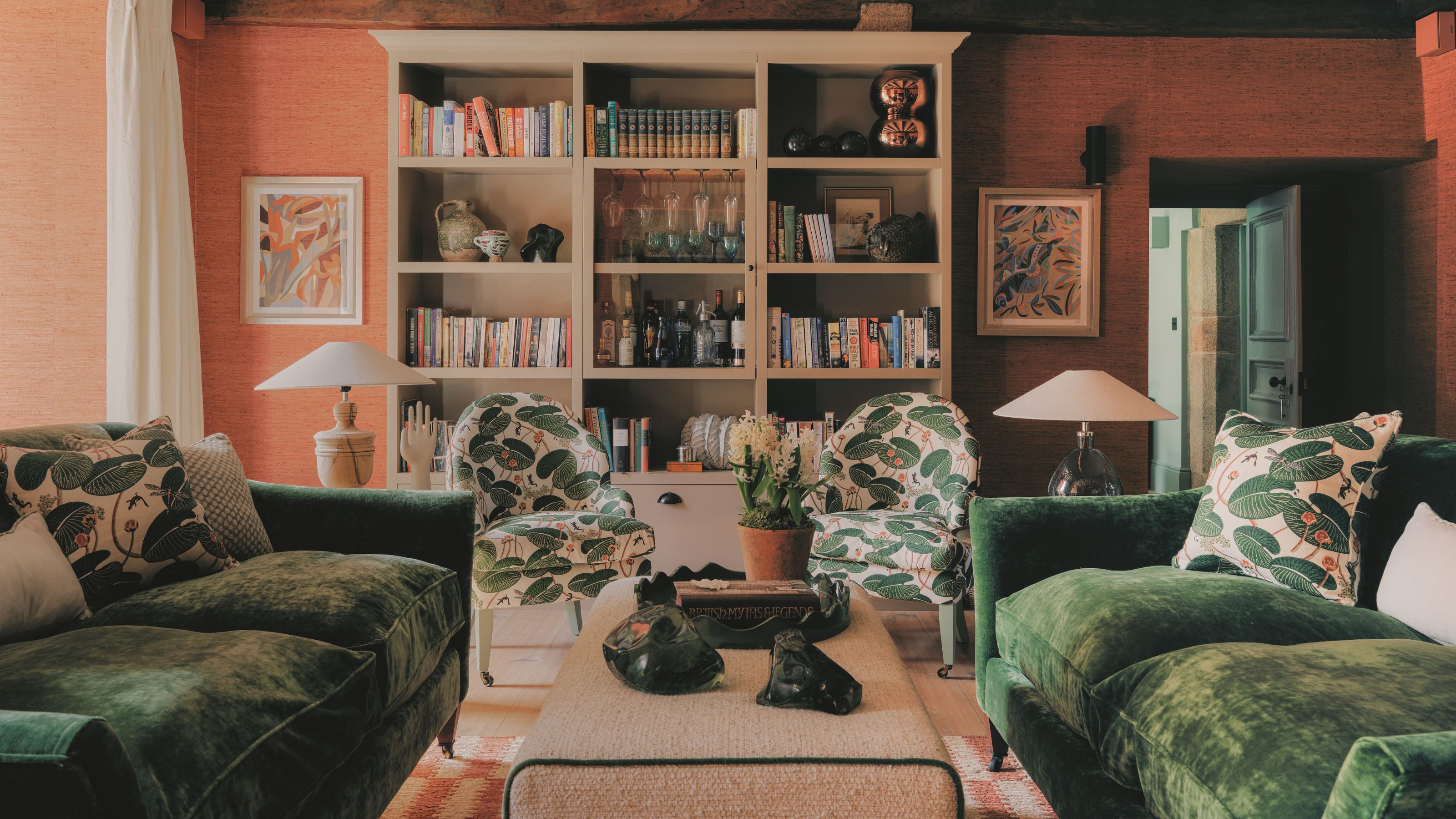 Gemütliches Wohnzimmer mit Terrakottawänden, dunkelgrünen Samtsofas, Sesseln mit botanischem Muster und eingebauten Bücherregalen voller Bücher und Glaswaren.