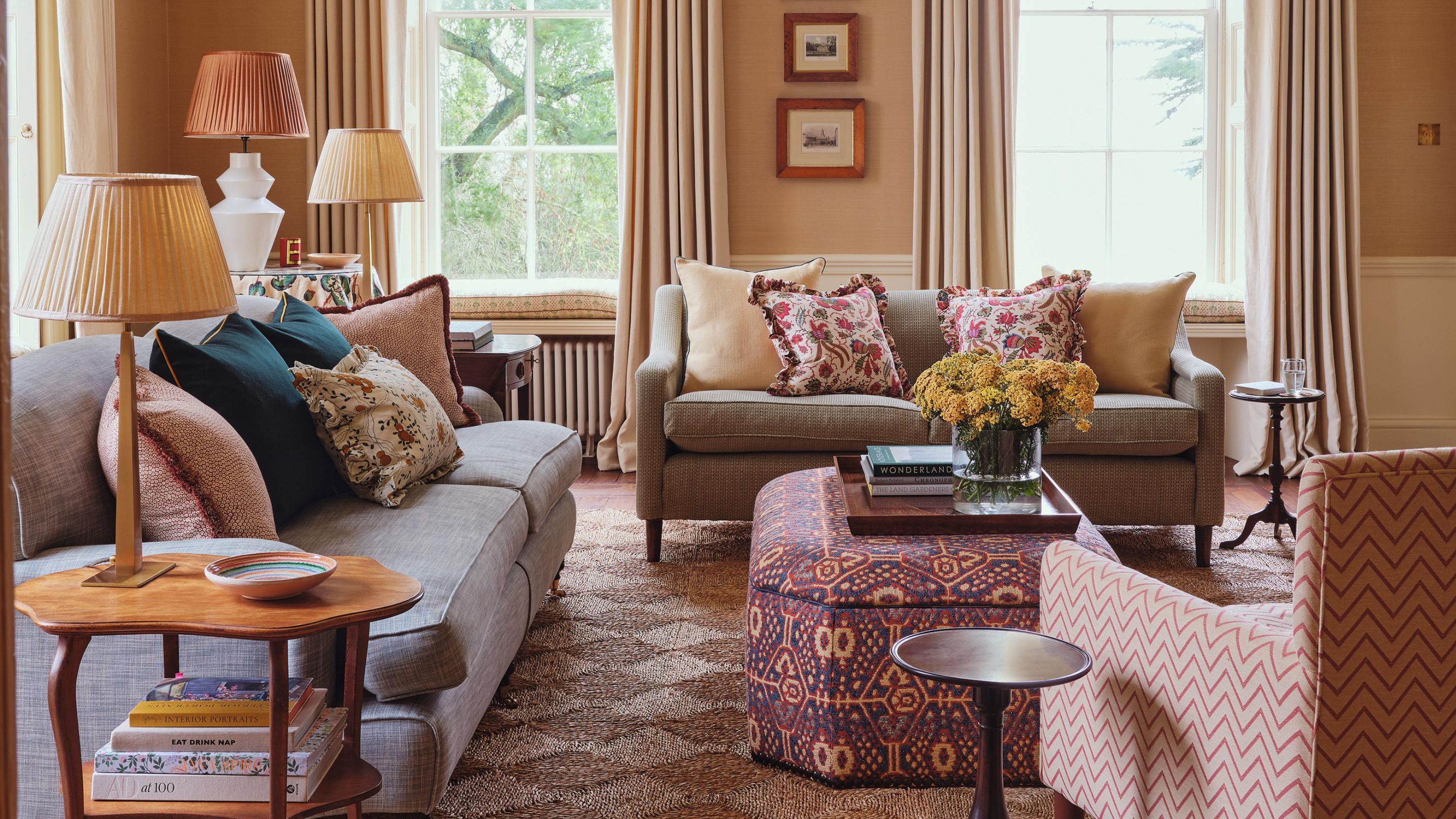 Ein Wohnzimmer im englischen Landhausstil mit einem gewebten Teppich, zwei neutralen Sofas, gemusterten Kissen, einem gemusterten Ottoman und einem gemusterten Sessel