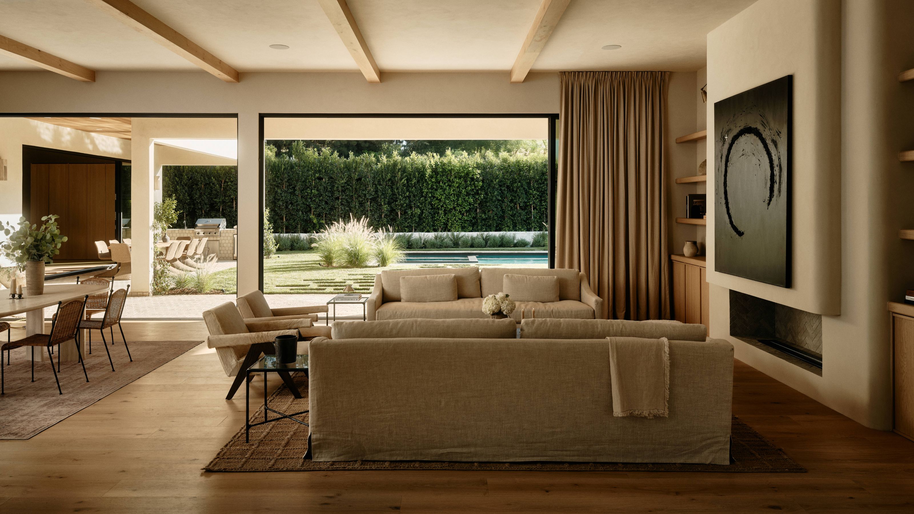ein neutrales, organisches, modernes Wohnzimmer und ein offener Essbereich mit großem Kamin und Leinensofas, der zu einer Poolterrasse und einer Außenküche führt