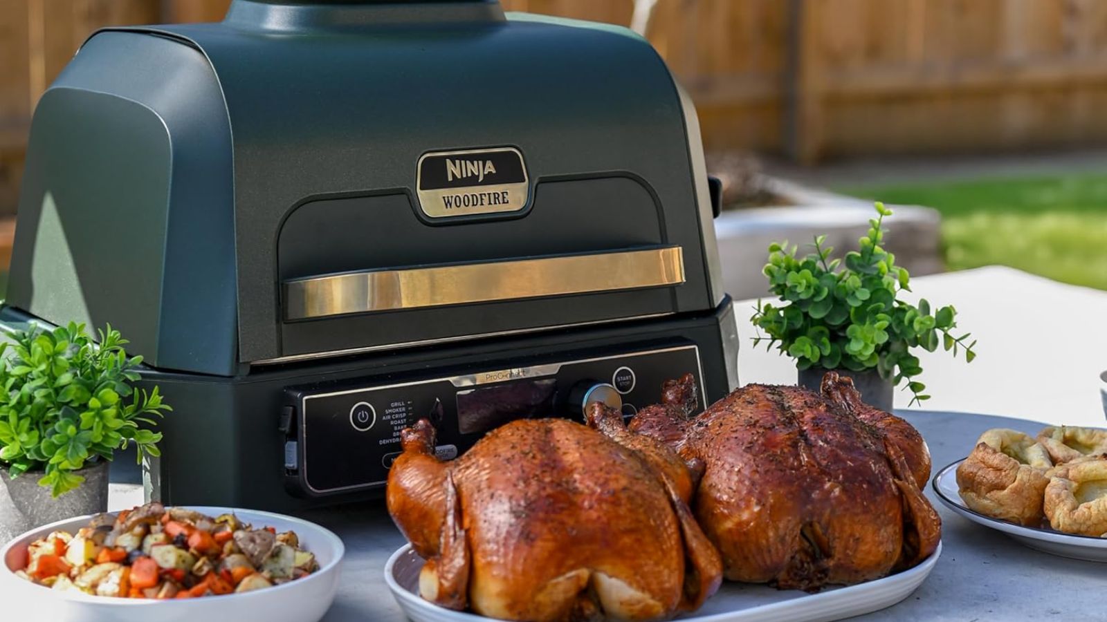 Der blau-goldene Ninja Woodfire Pro-Elektrogrill auf einem Tisch im Freien, umgeben von Brathähnchen, Beilagen und Kartoffeln 