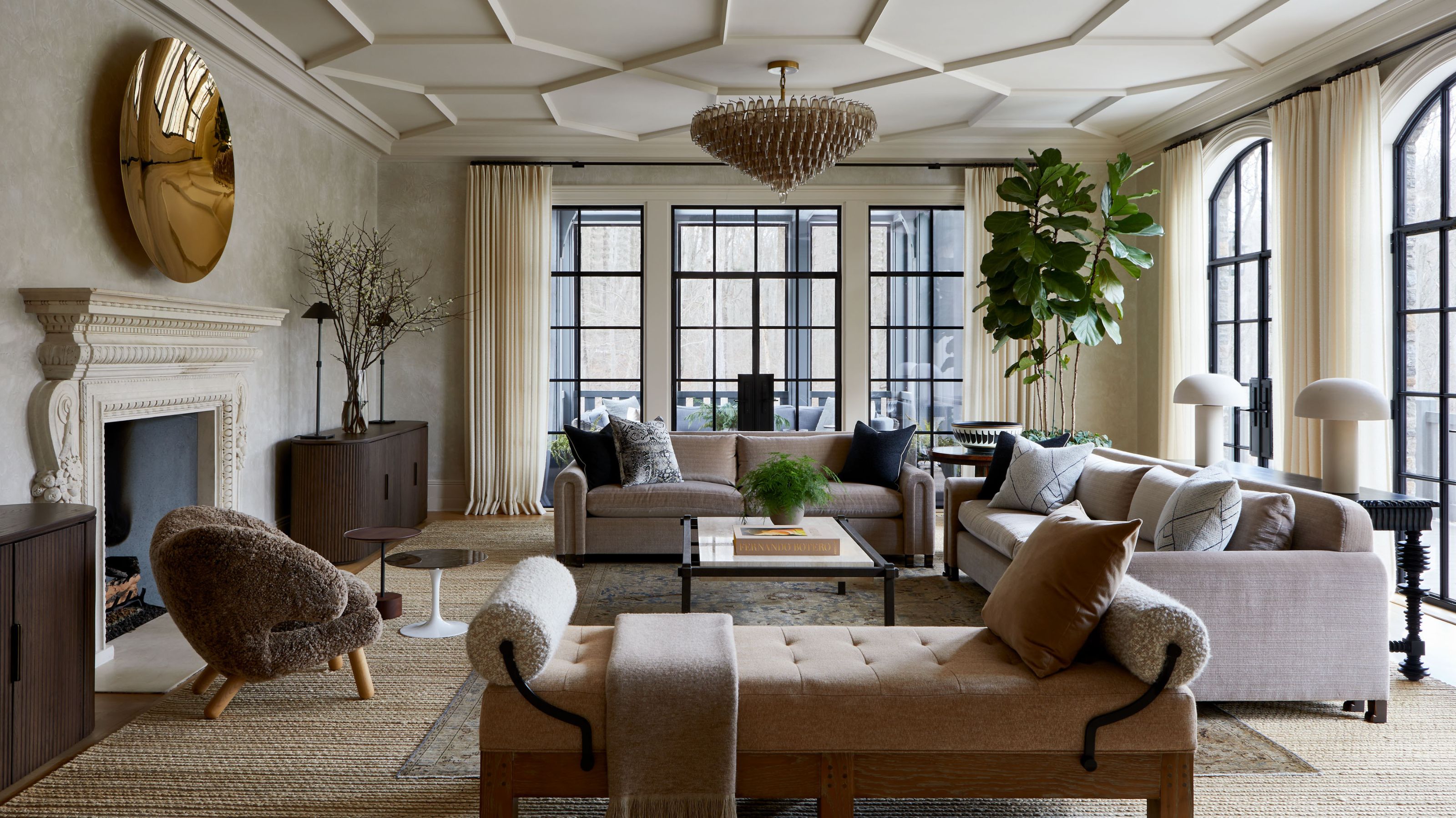 Ein großes Wohnzimmer mit dekorativem Deckenputz, einer neutralen Farbpalette und einer Wand aus Glastüren, die zu einer Veranda an zwei Wänden führen. Zwei taupefarbene Sofas mit Kissen und eine beige Chaiselongue. 