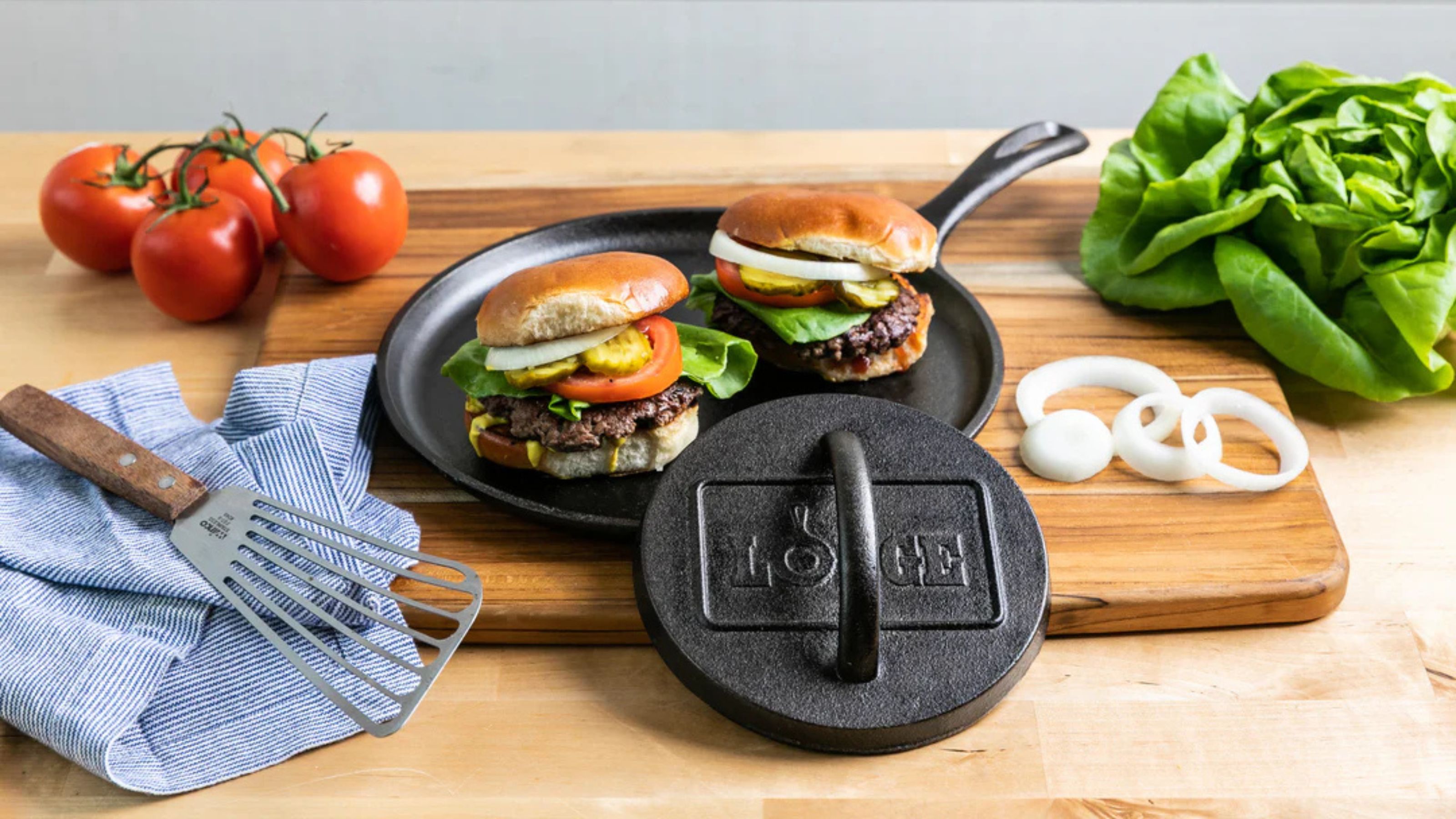 Eine gusseiserne Burgerpresse von Lodge lehnt an einem Teller mit zwei Burgern