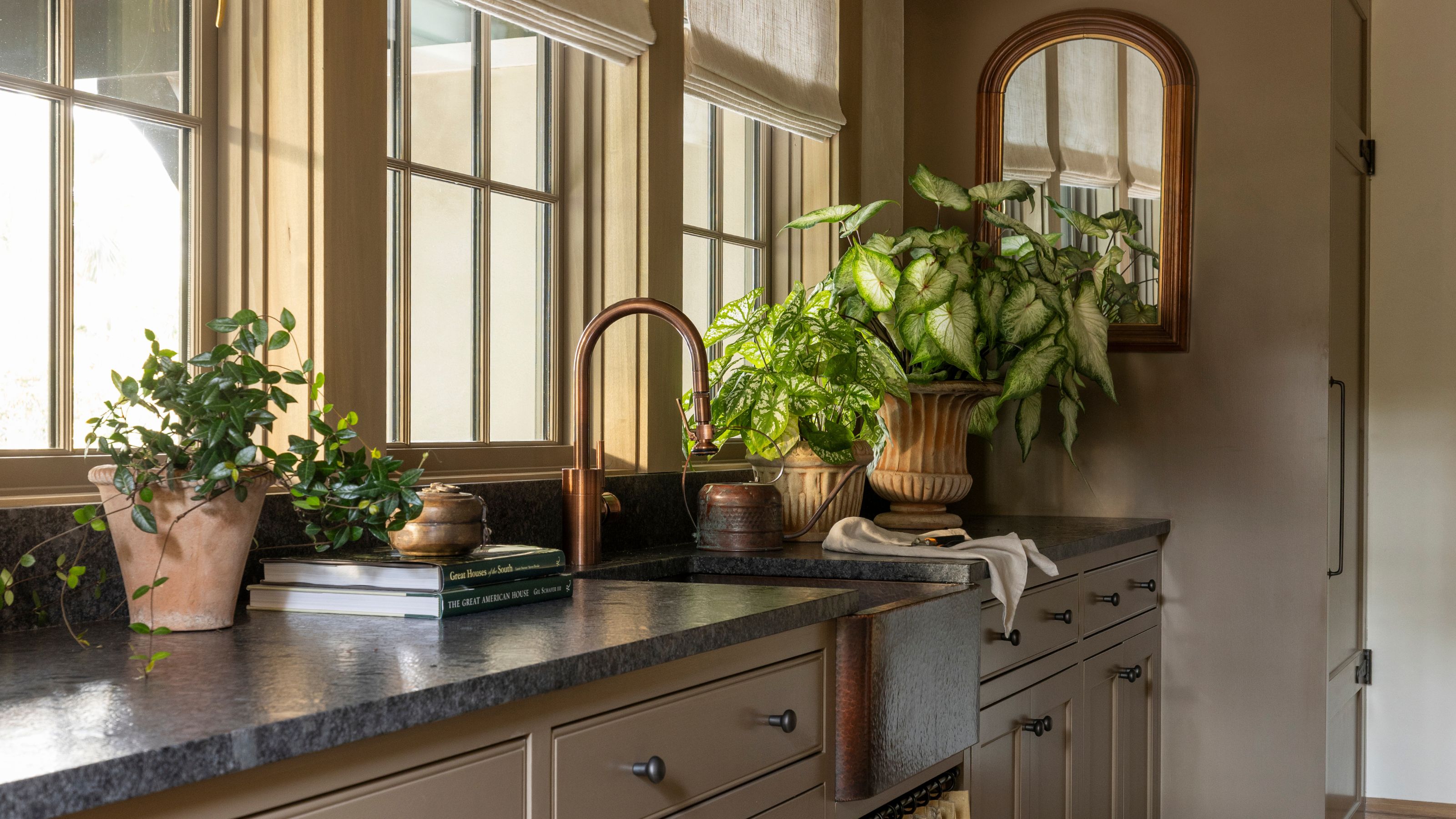 Mudroom mit Schränken und Wänden in Farrow & Ball's Broccoli braun, drei große Fenster, eine Arbeitsplatte aus dunklem Marmor, ein Waschbecken mit einem Schrank mit Schürze darunter und Pflanzen in rustikalen Töpfen.