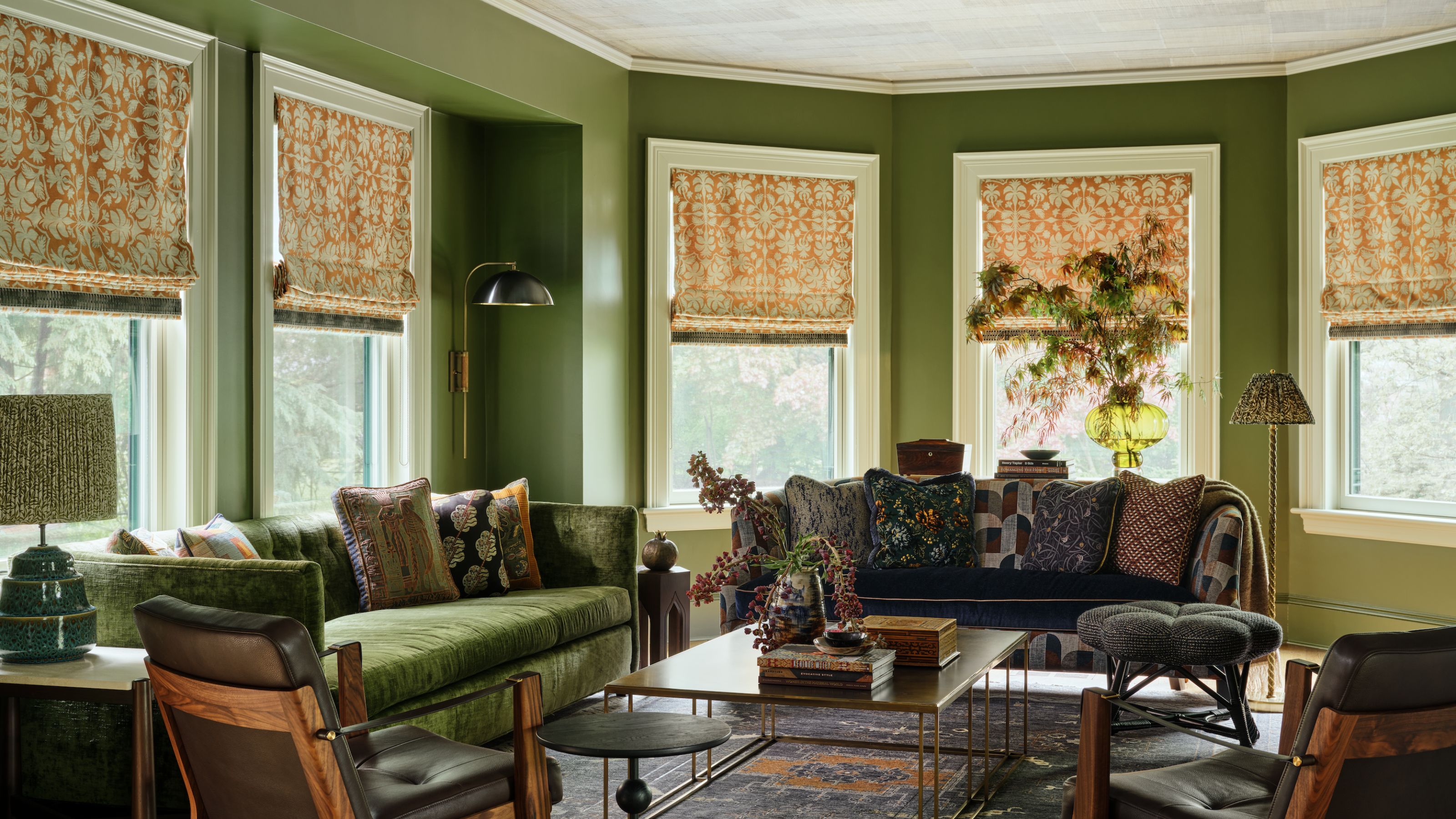 Ein gemütliches Wohnzimmer mit moosgrünen Wänden, fünf Fenstern mit orange gemusterten Jalousien, weißen Verzierungen und Decken, zwei Samtsofas und zwei passenden Leder- und Holzsesseln