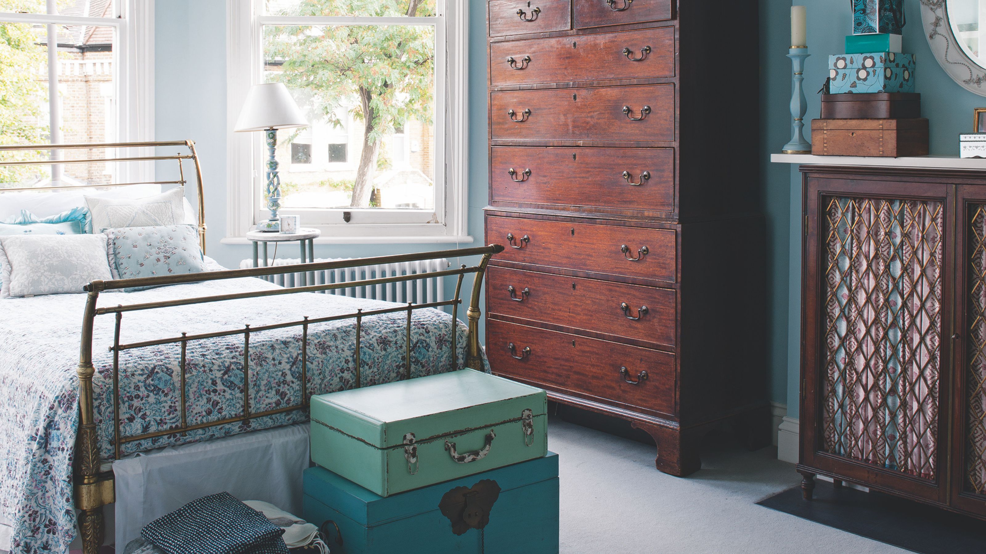 Blaues Gästezimmer mit goldenem Bett, Kommode und Schrank aus dunklem Holz, am Fußende des Bettes gestapelten blauen Koffern und großen Erkerfenstern.