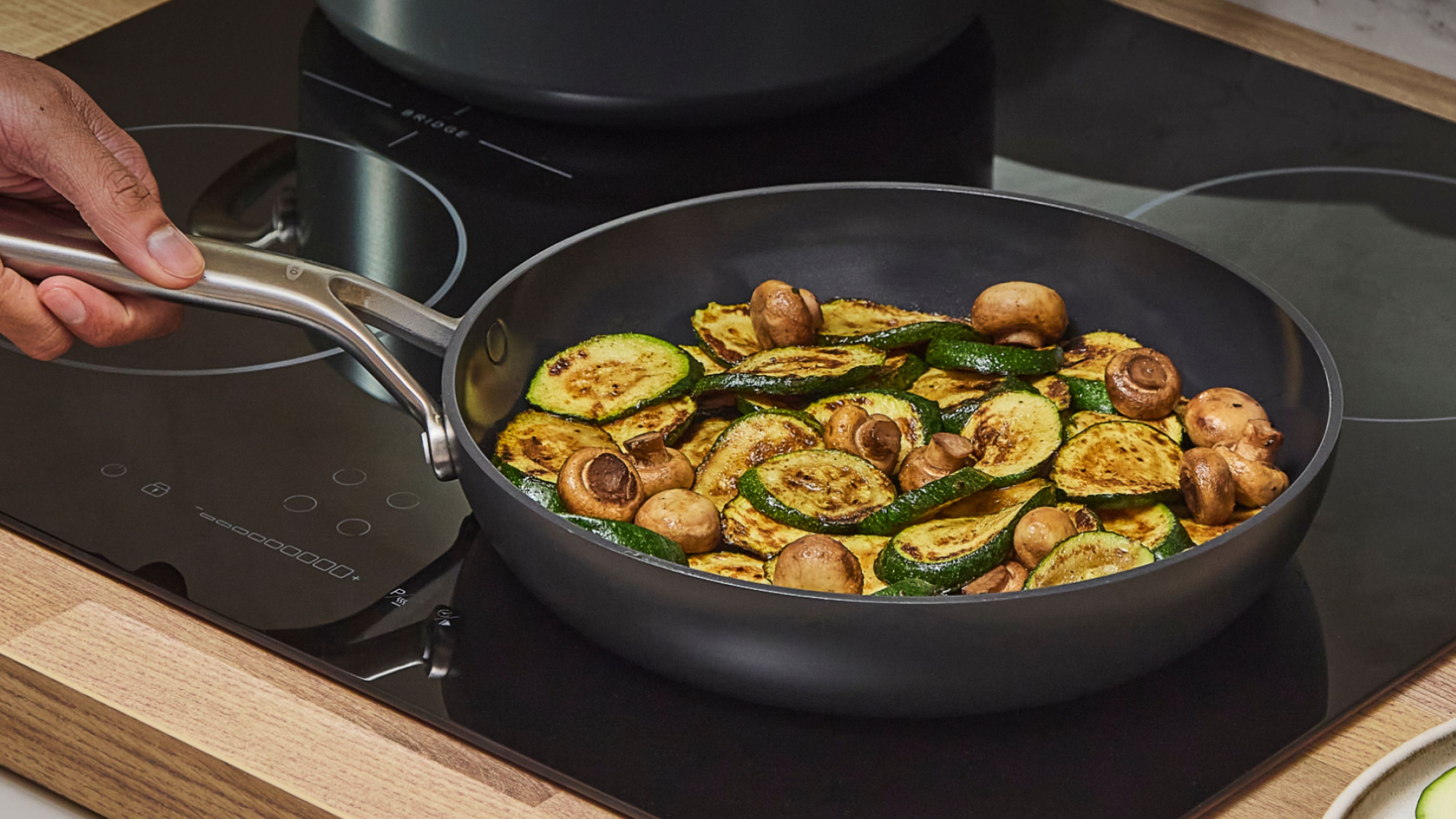 Eine schwarze Keramikpfanne auf einem schwarzen Herd mit Pilzen und Zucchini darin. 