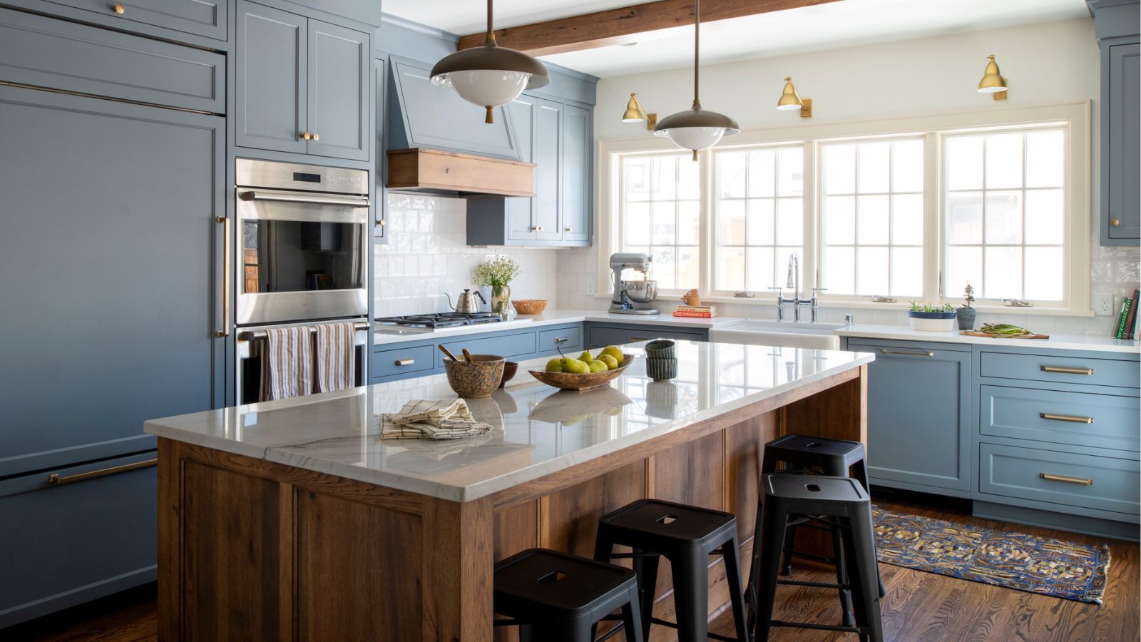 A transitional kitchen with blue-gray cabinets, a wooden island with white countertops, black bar stools, and wood ceiling beams. Brass wall sconces by the window and a butler's sink.